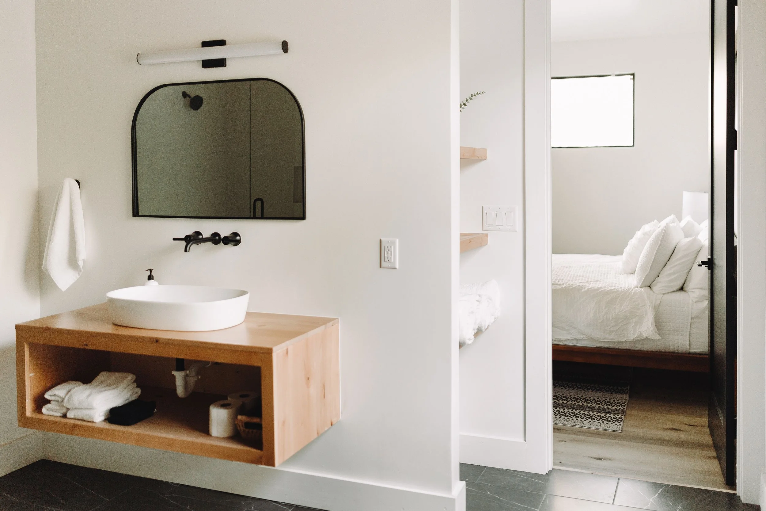 A modern bathroom with a floating wooden vanity with a white vessel sink, black fixtures, and a rounded mirror. A white towel hangs on a hook beside the vanity. There are two floating shelves on the wall beside the door, holding folded towels and a s