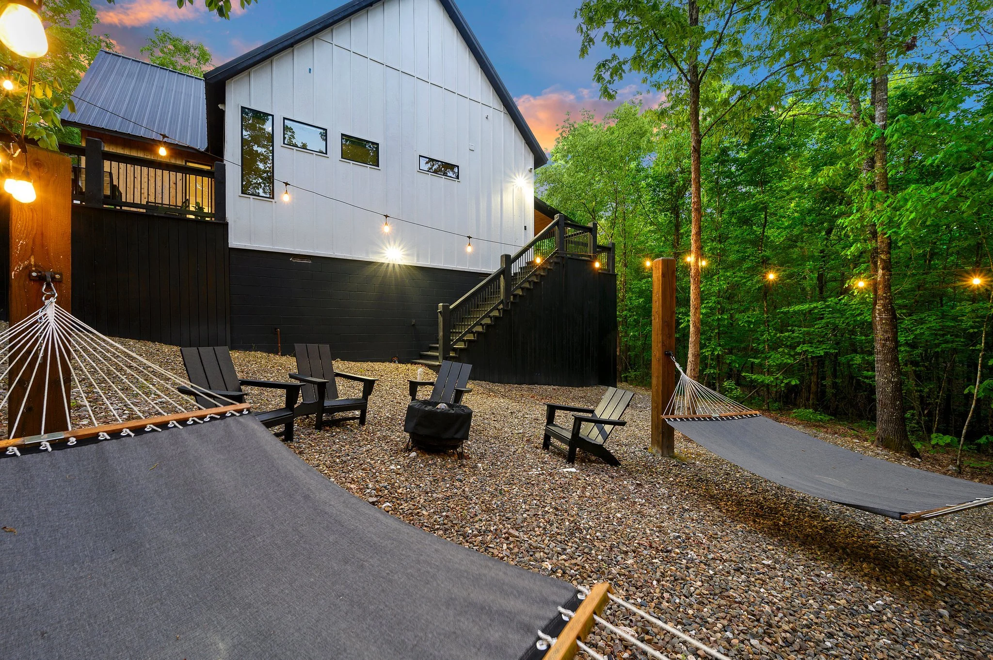 Outdoor backyard with string lights, black and white house, gravel ground, black chairs, hammocks, and green trees.
