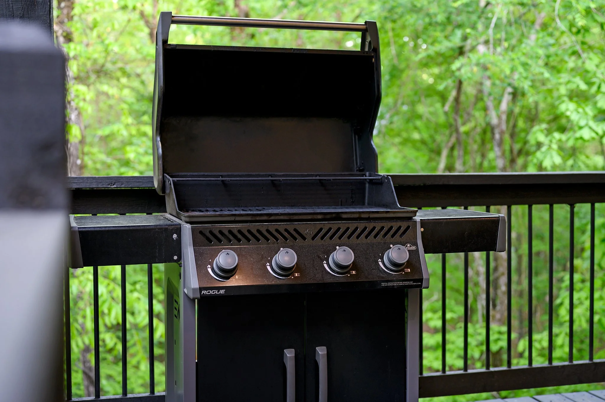 Black outdoor gas grill with four control knobs, open lid, and surrounded by green trees in the background on a wooden deck.