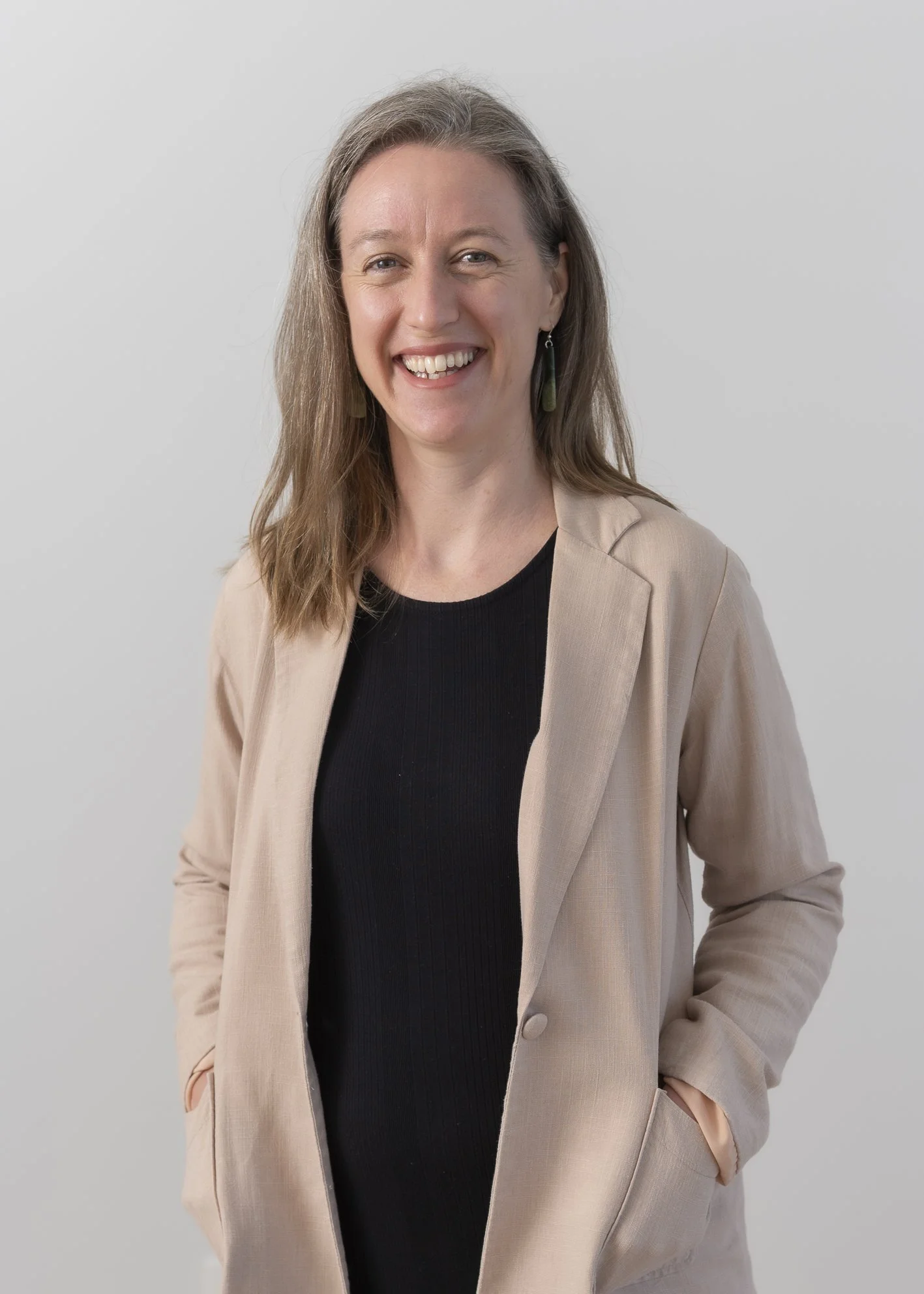 Portrait of a woman with long, light brown hair, smiling, wearing a beige blazer over a black top, standing against a plain gray background.