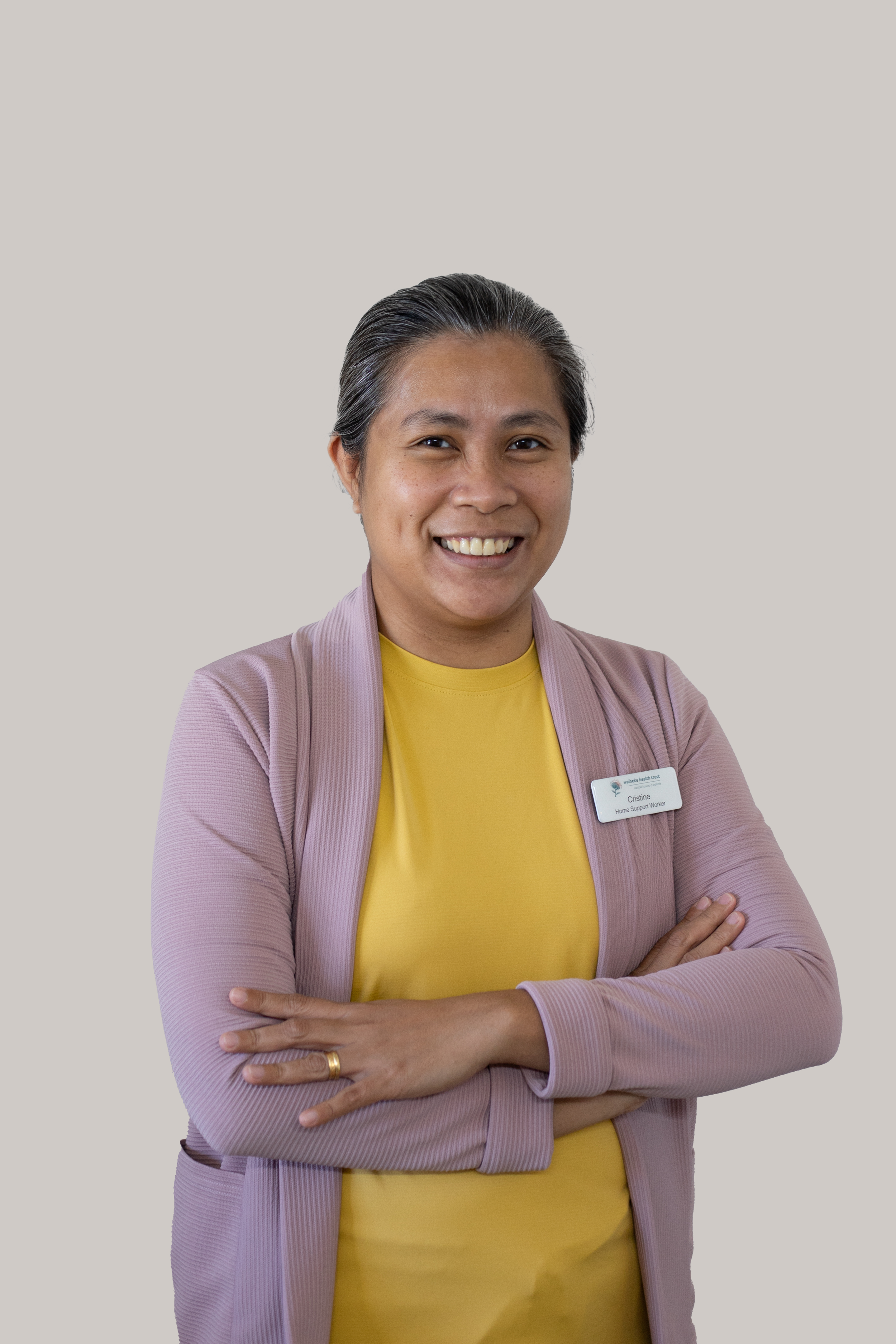 A woman with short dark hair smiling, wearing a yellow shirt and a lavender blazer, with arms crossed, in front of a plain light gray background.
