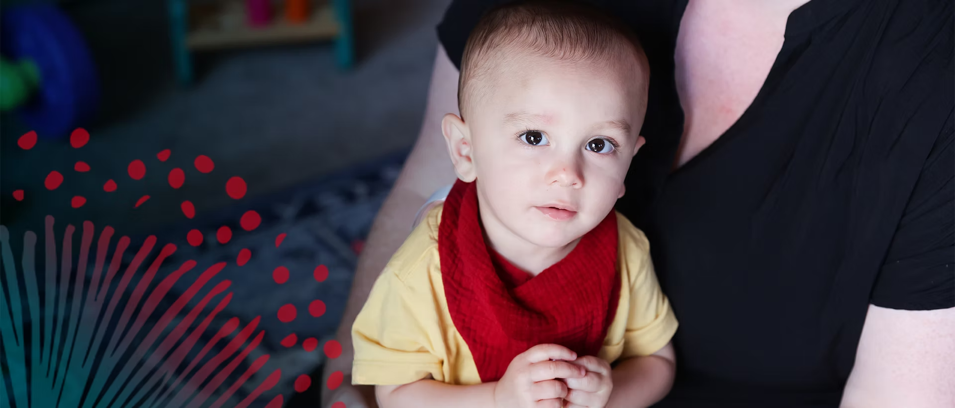 A young child with short light brown hair and blue eyes, wearing a yellow shirt and a red bib, is being held by an adult wearing a black shirt. The child is looking into the camera with a slight smile.