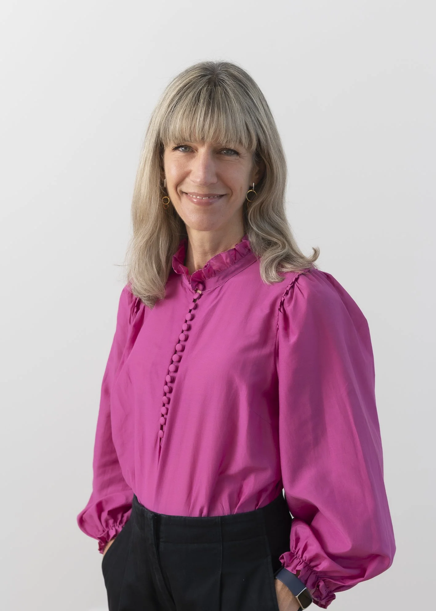 A woman in a pink blouse with ruffled collar and button details, wearing black pants and a smartwatch, standing against a plain white background.