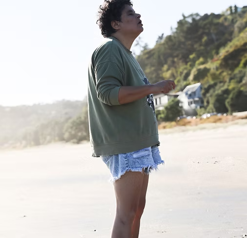 Person standing on a beach with hills and trees in the background, wearing a green long-sleeve shirt and denim shorts.