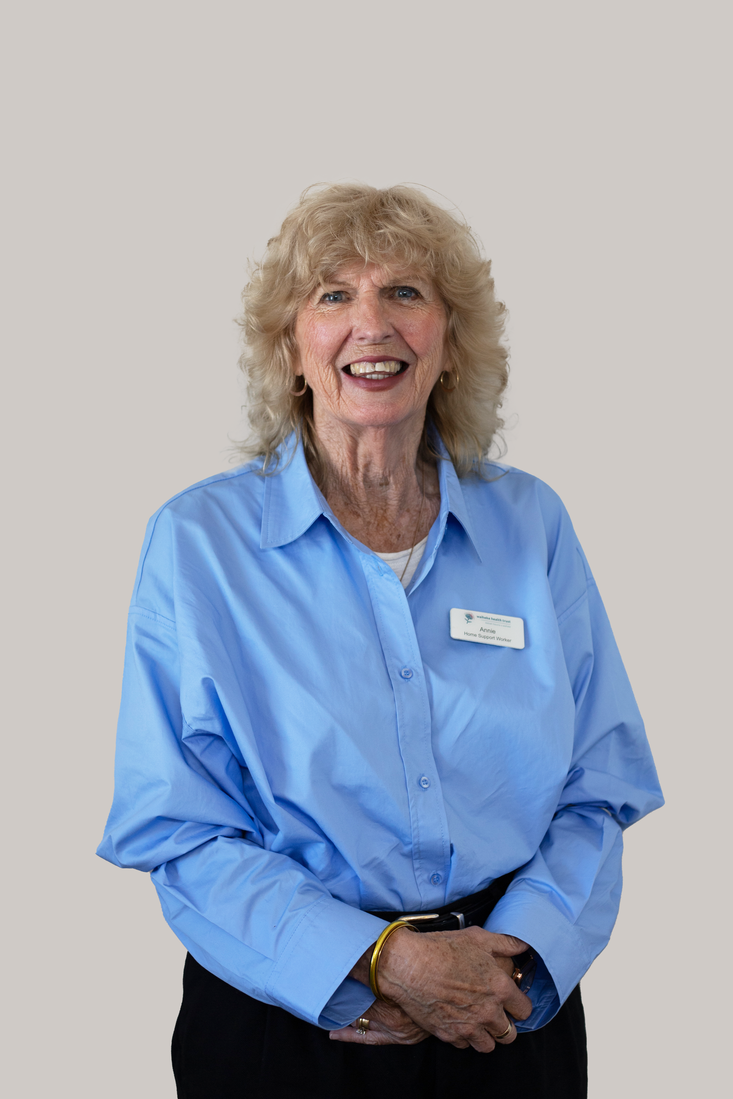An elderly woman with curly blonde hair, wearing a light blue collared shirt and a name badge, smiling and standing against a plain background.