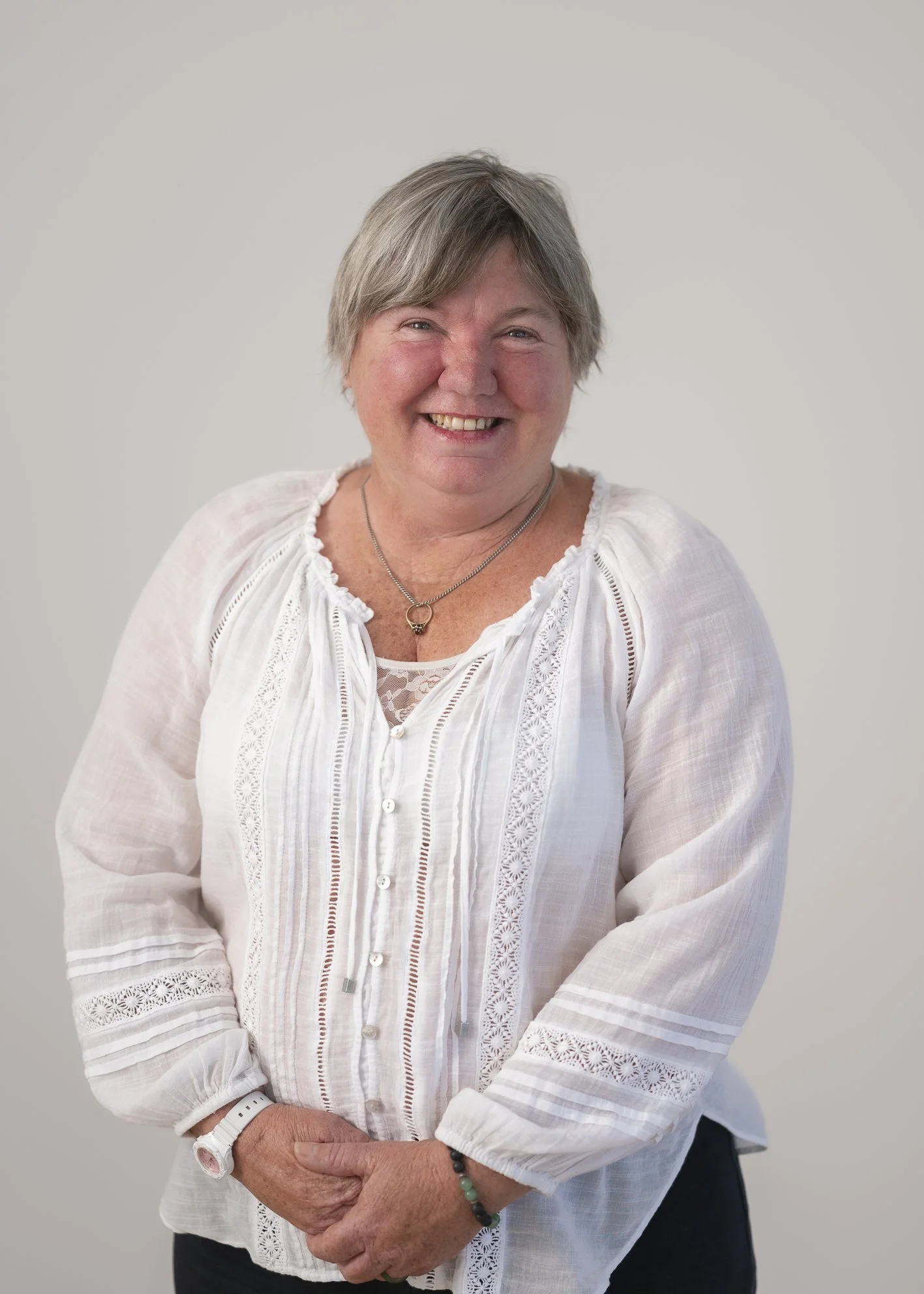 A smiling older woman with short gray hair wearing a white blouse with lace details, a necklace, a white watch, and a bracelet, standing against a plain light-colored background.