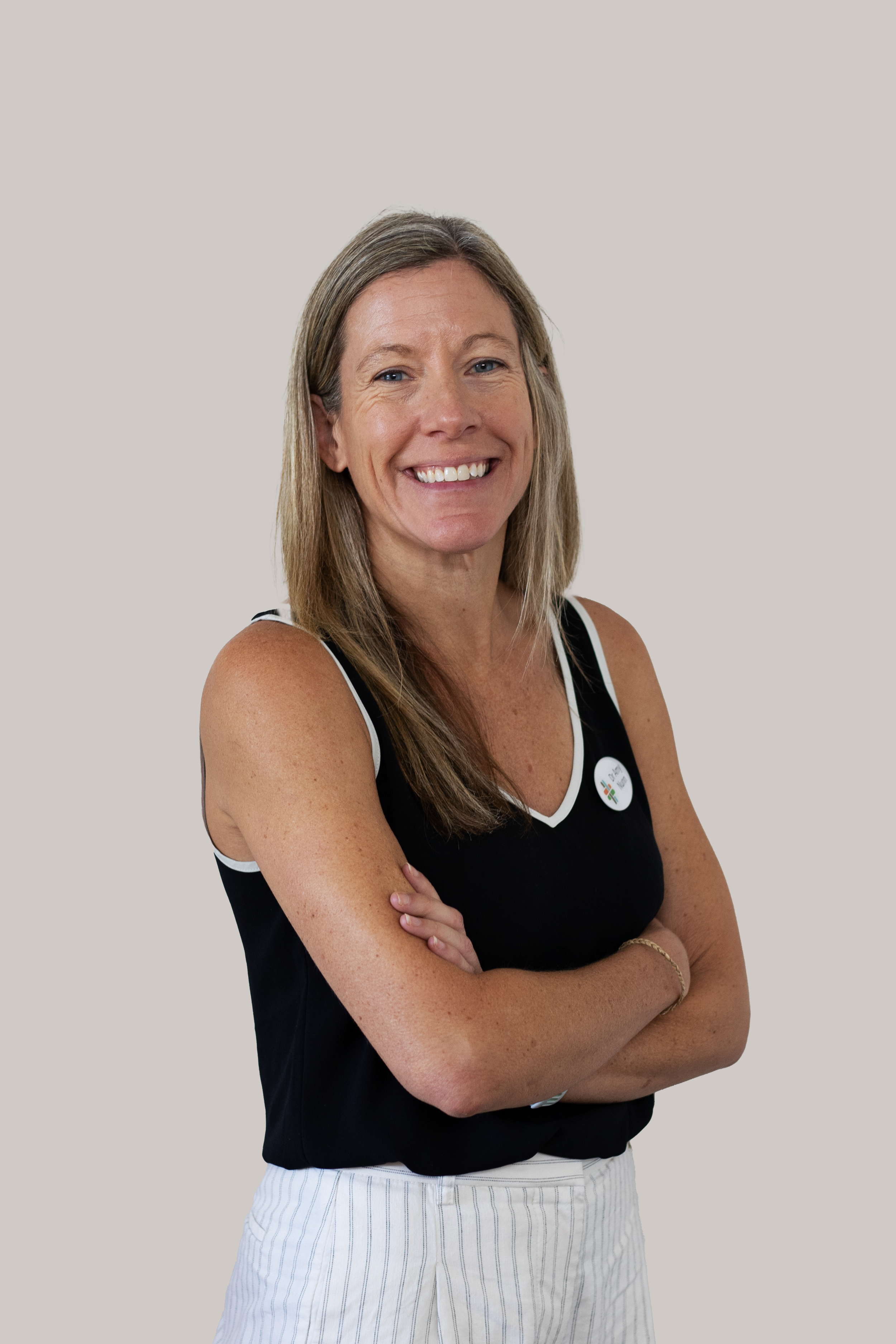 A woman with long light brown hair, wearing a black sleeveless top with white trim, standing with arms crossed and smiling, against a plain light gray background.
