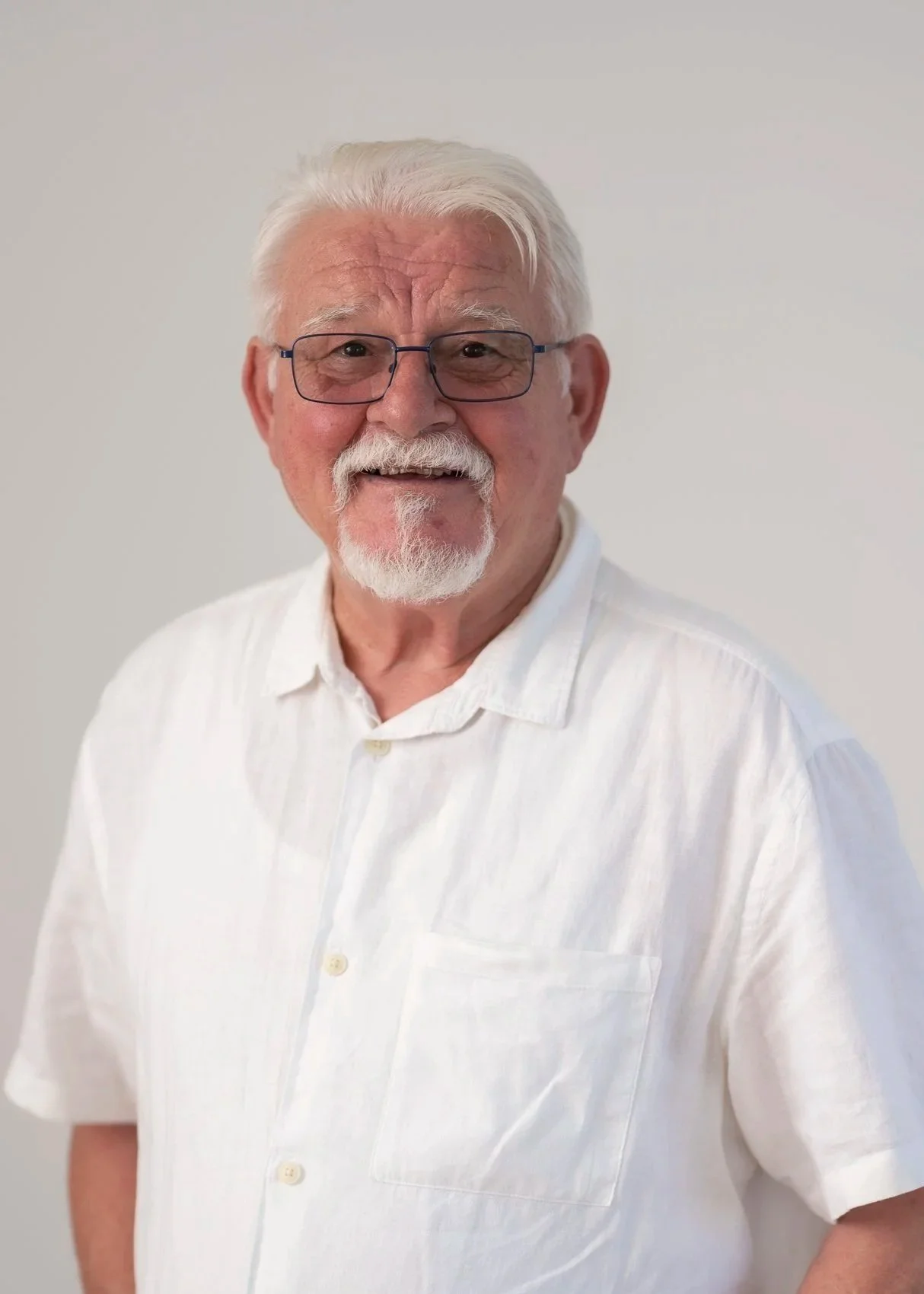 An elderly man with white hair, glasses, a mustache, and a goatee, smiling and wearing a white button-up shirt against a plain white background.