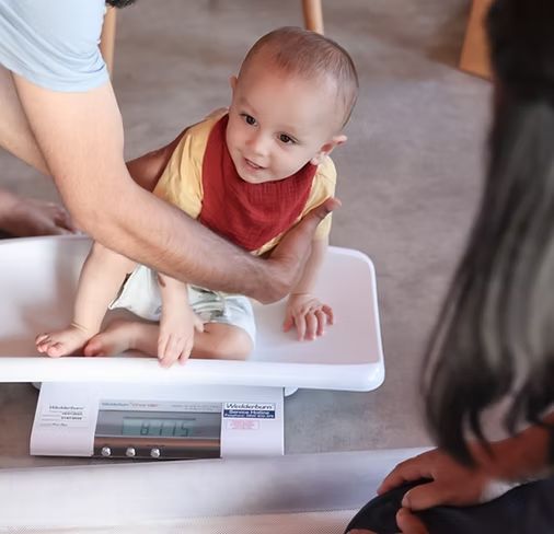 A baby sitting on a digital scale, being weighed by a nurse. The baby is wearing a yellow shirt and a red bib, while a woman on the right watches.