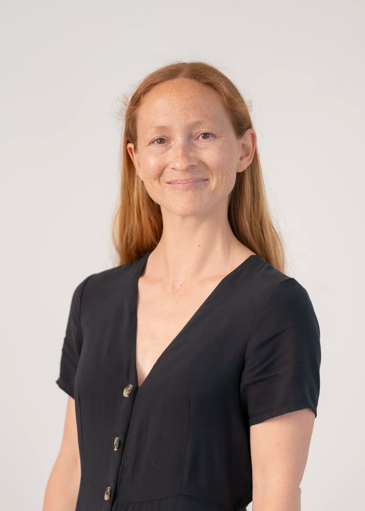 A woman with red hair wearing a black dress standing against a plain white background.