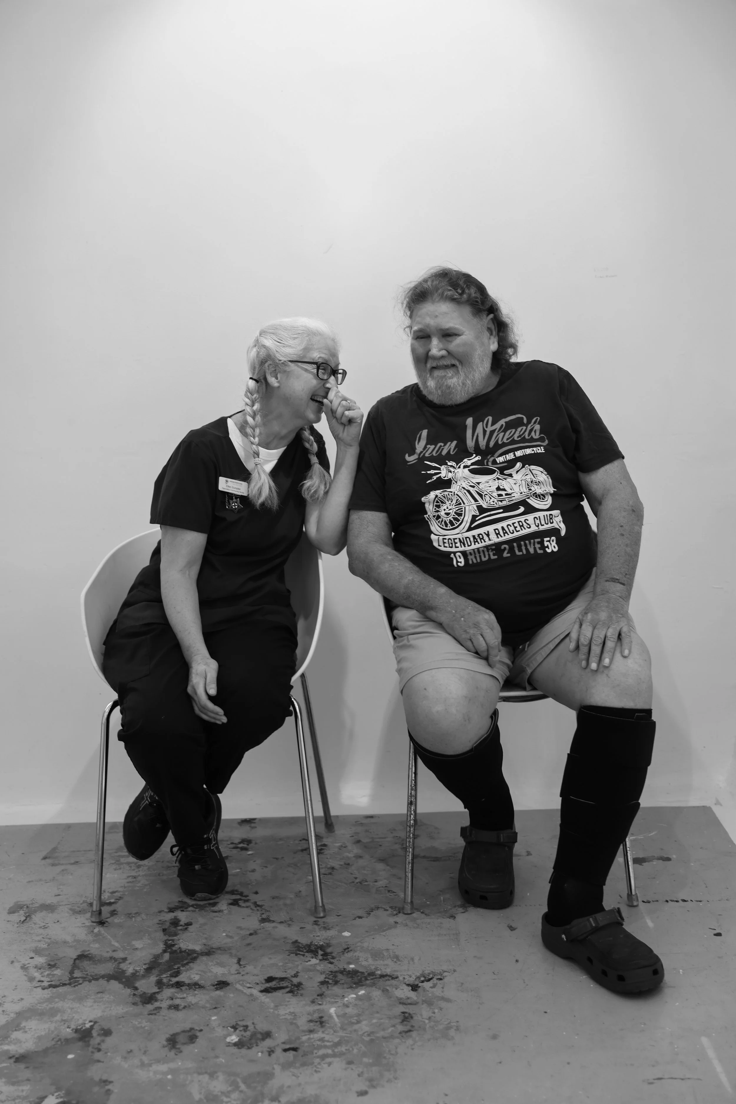 A woman and a man are sitting on chairs against a plain wall, laughing and sharing a moment of joy. The woman is wearing glasses, has long hair in braids, and is dressed in a dark uniform. The man is wearing a T-shirt with a motorcycle graphic, shorts, and black boots, and has curly hair and a beard.