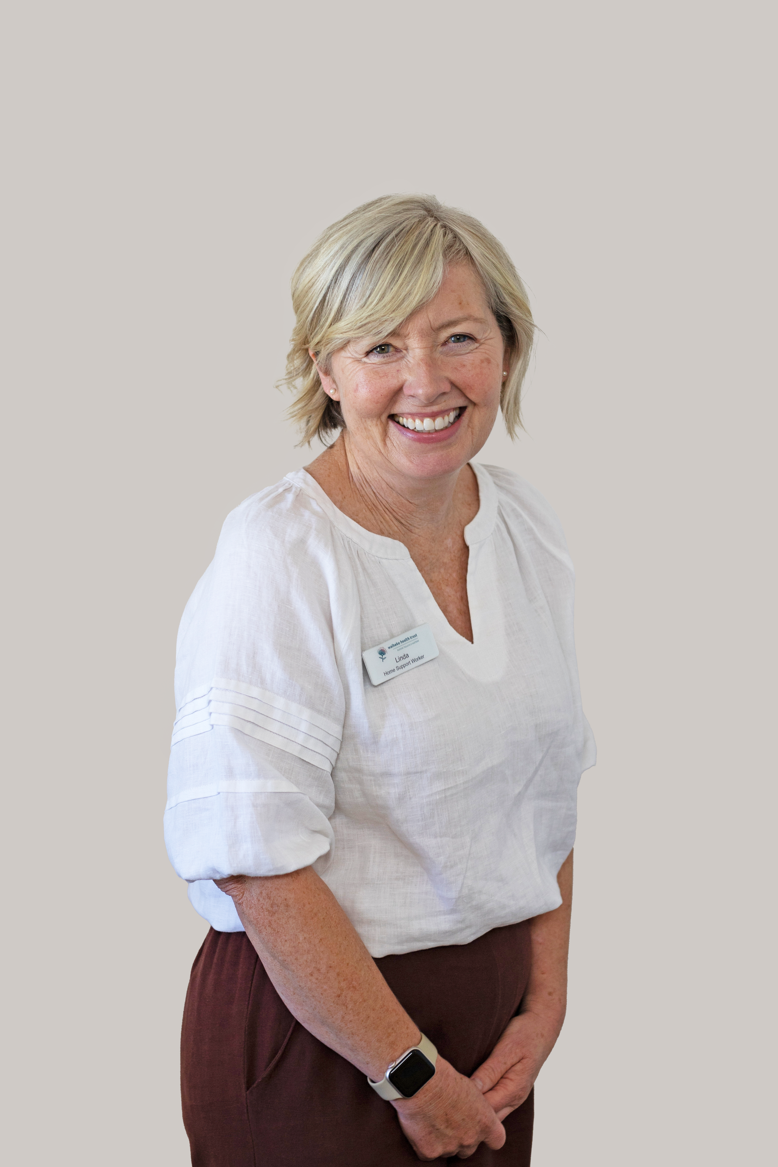 A smiling blonde woman wearing a white blouse, brown pants, & a smartwatch on her left wrist, standing against a plain light background.