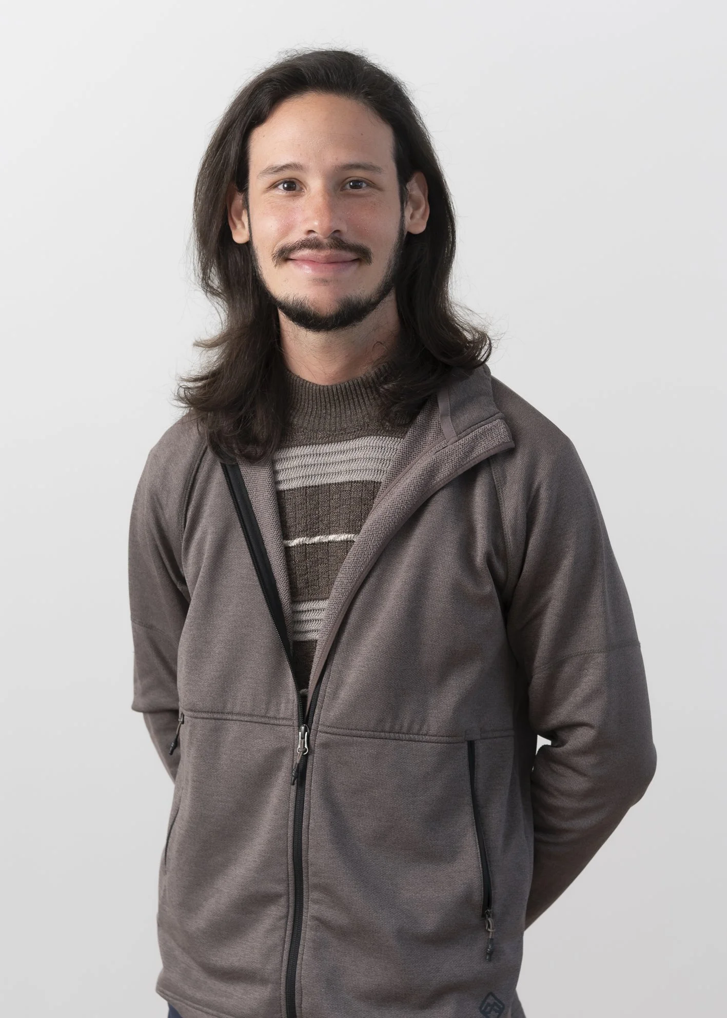 A man with long hair, a beard, and a mustache, wearing a brown jacket and a striped sweater, standing against a plain light background.