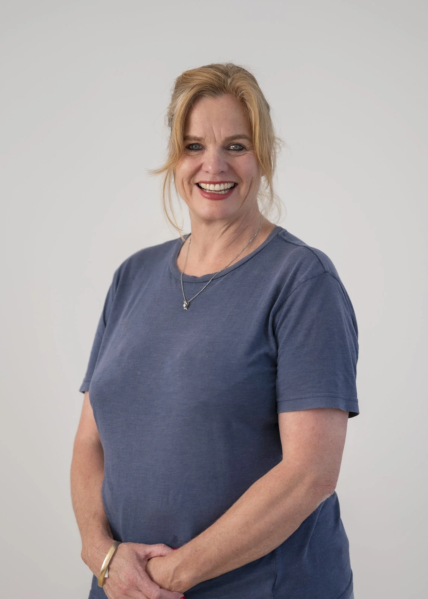 A woman with blonde hair styled in a loose updo, wearing a blue T-shirt and a silver necklace, standing against a plain light-colored background, smiling at the camera.
