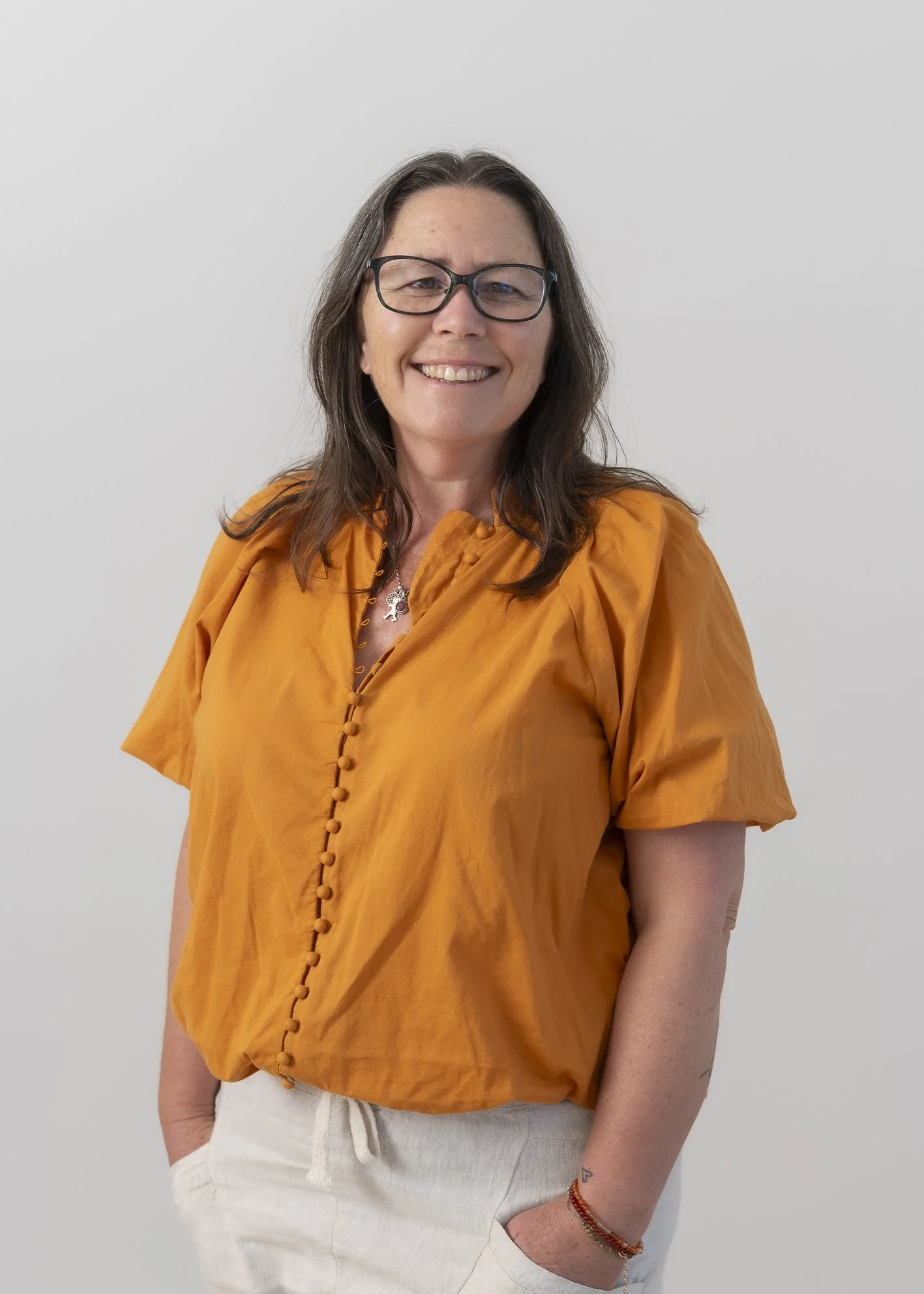 A woman with long brown hair, black glasses, wearing an orange blouse, white pants, and a bracelet, standing against a plain white wall, smiling at the camera.