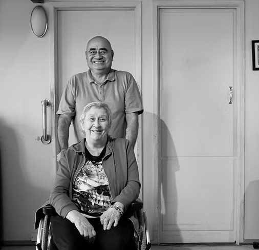 A smiling elderly woman in a wheelchair with a smiling man standing behind her in a room with plain walls and a door.