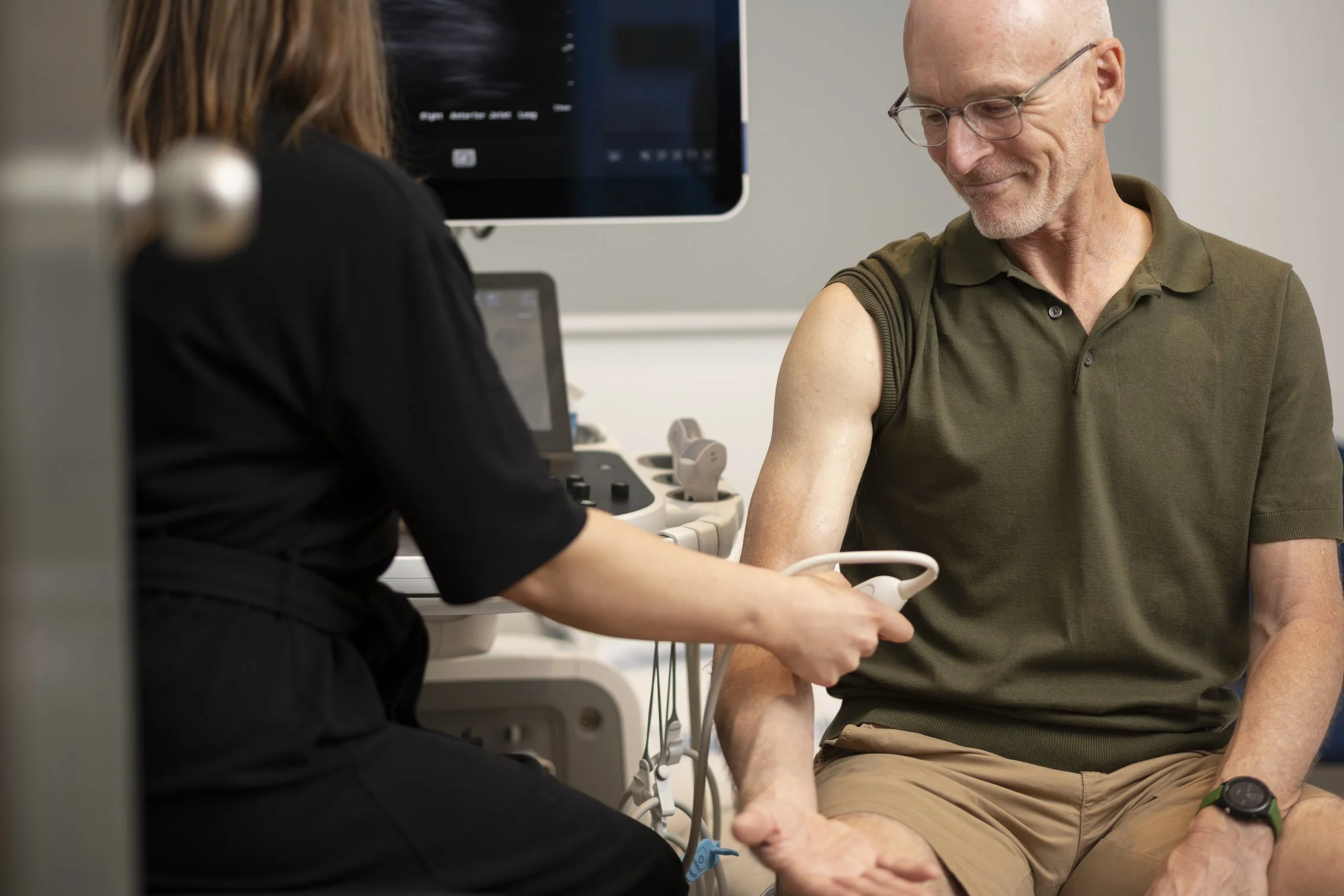 A man sits in a medical clinic preparing for an X-ray, smiling while a healthcare professional assists him with the procedure.