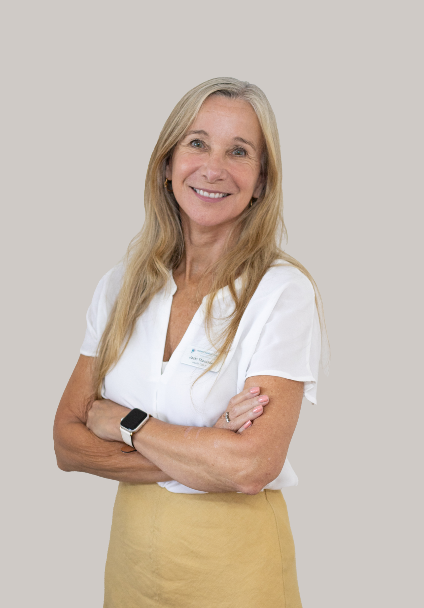 A smiling middle-aged woman with long blonde hair, wearing a white short-sleeved blouse, tan skirt, and a smartwatch, standing with her arms crossed against a plain light-colored background.