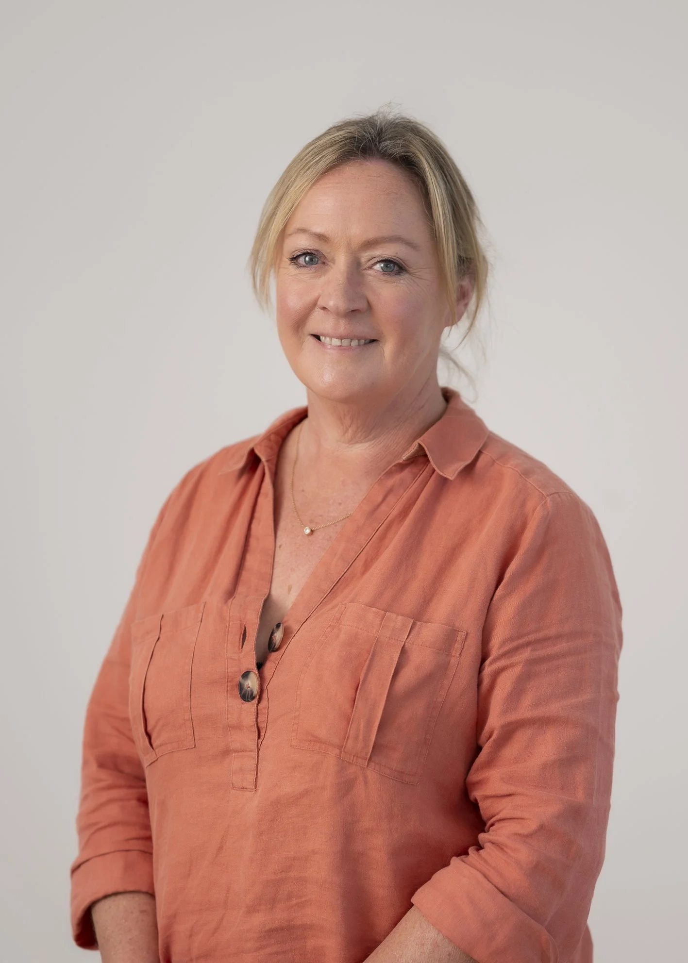 A woman with blonde hair, blue eyes, wearing an orange button-up shirt and a delicate necklace, standing against a plain light gray background.