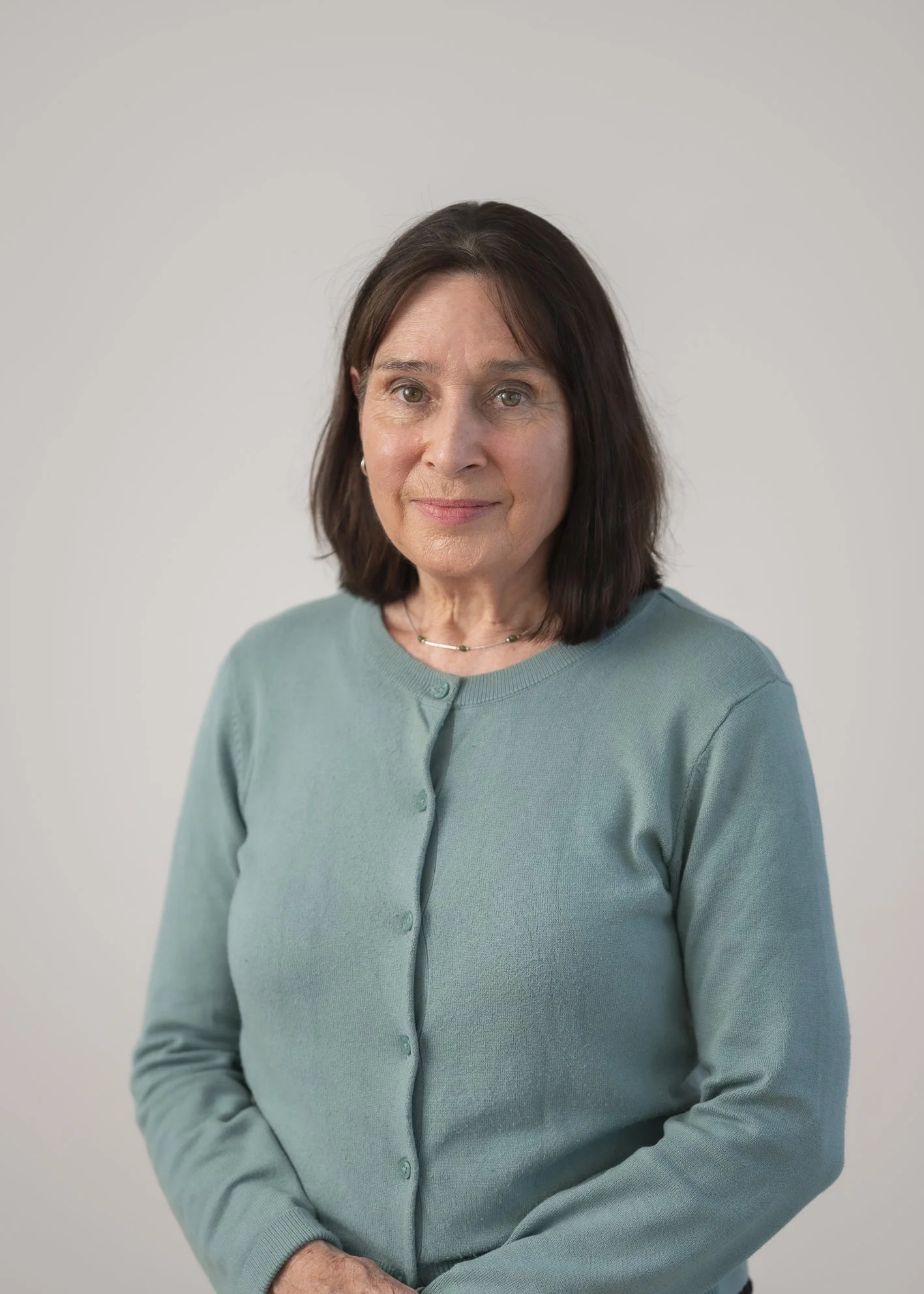 A portrait of a middle-aged woman with dark brown hair, wearing a light blue cardigan and a delicate necklace, standing against a plain gray background.