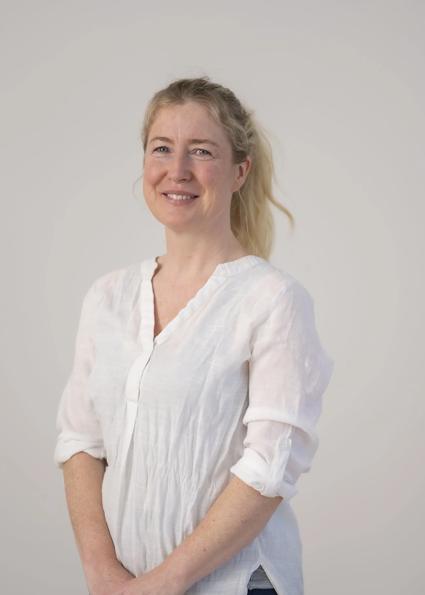 A woman with blonde hair tied in a ponytail, wearing a white blouse, smiling, against a plain light-colored background.