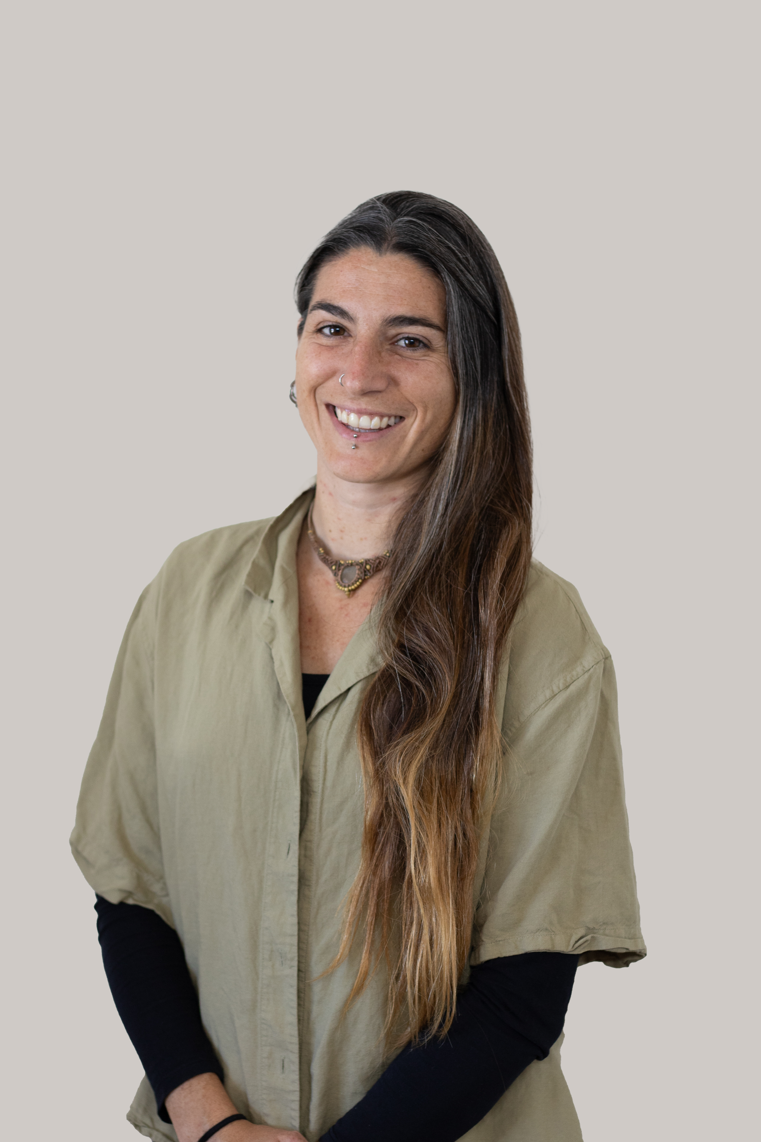 A woman with long, wavy brown hair, fair skin, and multiple facial piercings, smiling and looking at the camera, wearing a beige button-up shirt over a black long-sleeve shirt, accessorized with necklaces and a black wristband, against a plain light grey background.