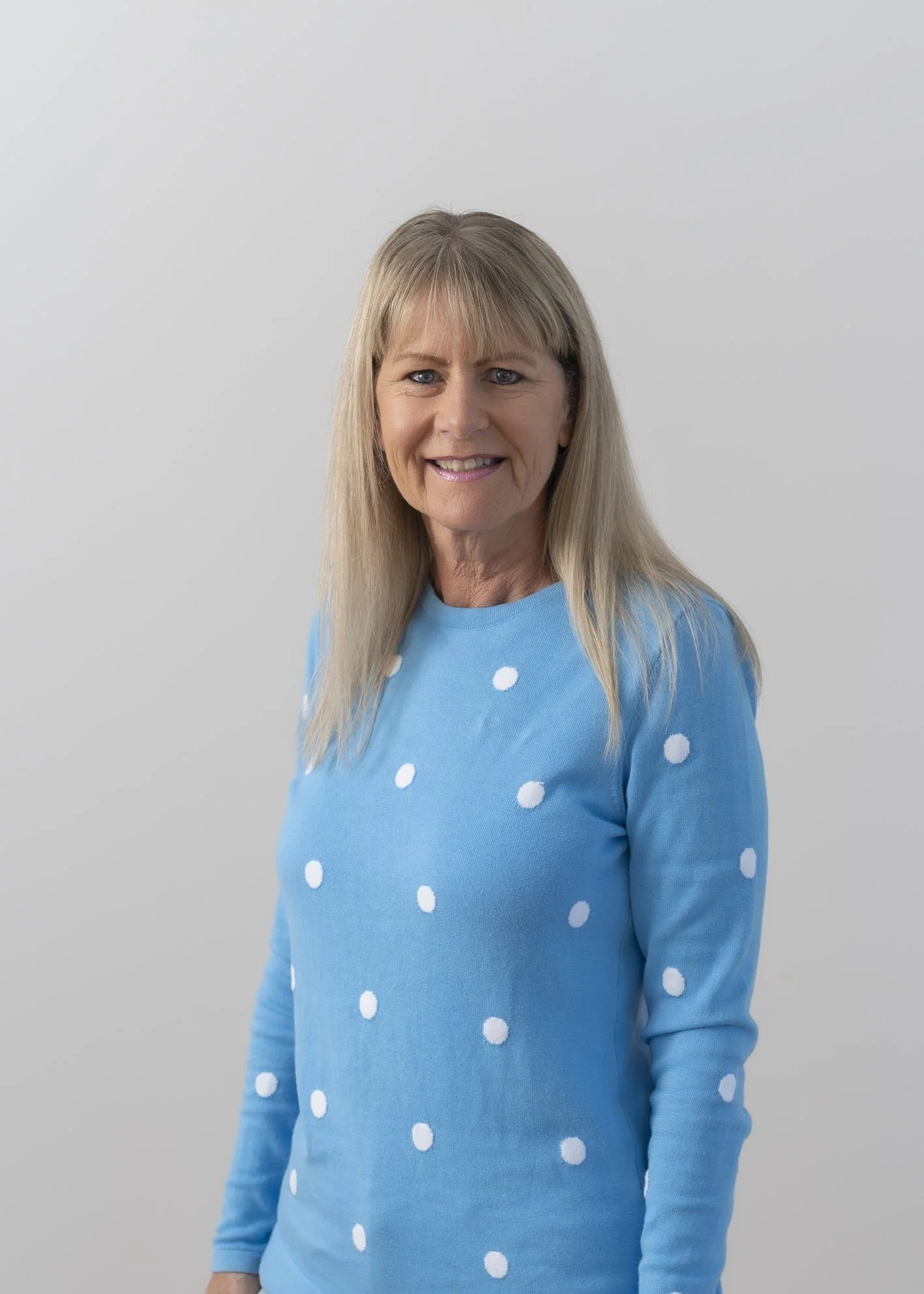 A middle-aged woman with long blonde hair wearing a blue sweater with white polka dots, smiling, against a plain light gray background.
