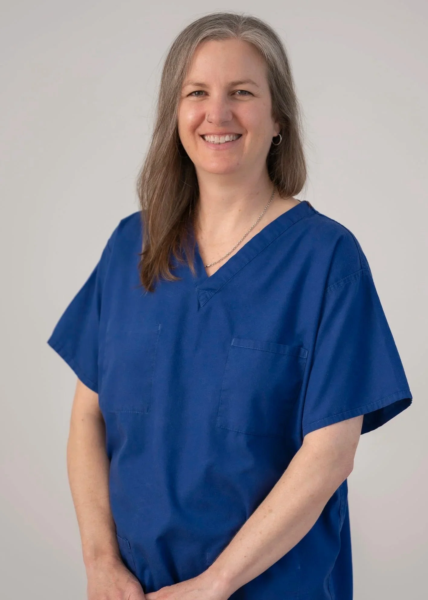 A woman wearing blue medical scrubs, smiling, short light brown hair, standing against a plain light gray background.