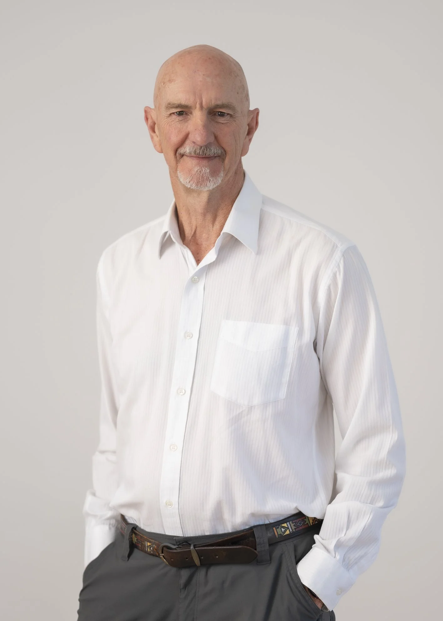 A senior man with a bald head and white beard wearing a white dress shirt and dark pants, standing against a plain light gray background.