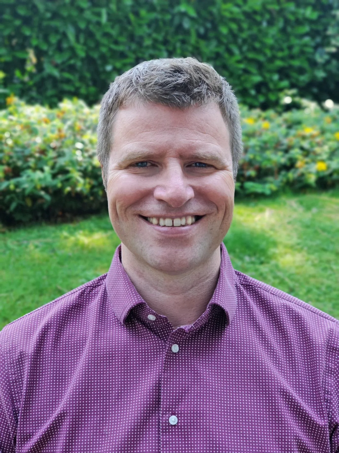 A man with short, light brown hair, blue eyes, and a broad smile, wearing a purple checkered shirt, standing outdoors in front of a green garden with bushes and trees.