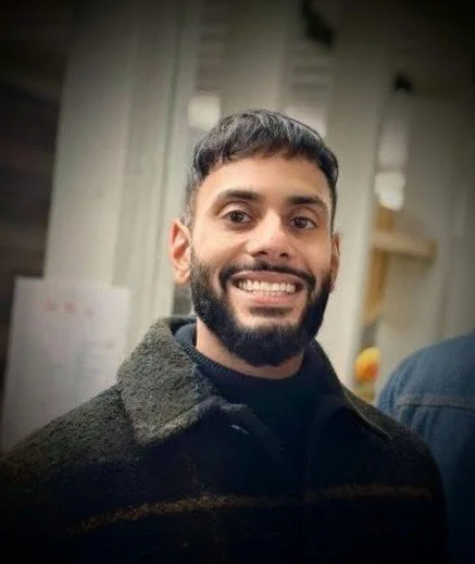 A man with dark hair and a beard smiling, wearing a dark coat, in an indoor setting.