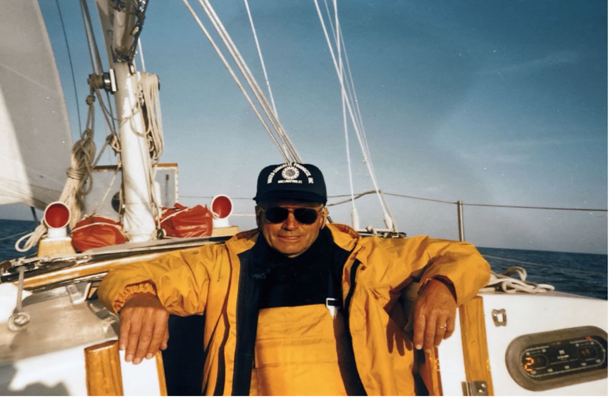 A man wearing sunglasses, a black cap, and a yellow jacket sitting on a sailboat in the open sea.