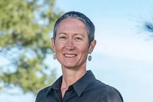 A middle-aged woman with short gray hair, wearing a dark button-up shirt, smiling outdoors with trees and a blue sky in the background.