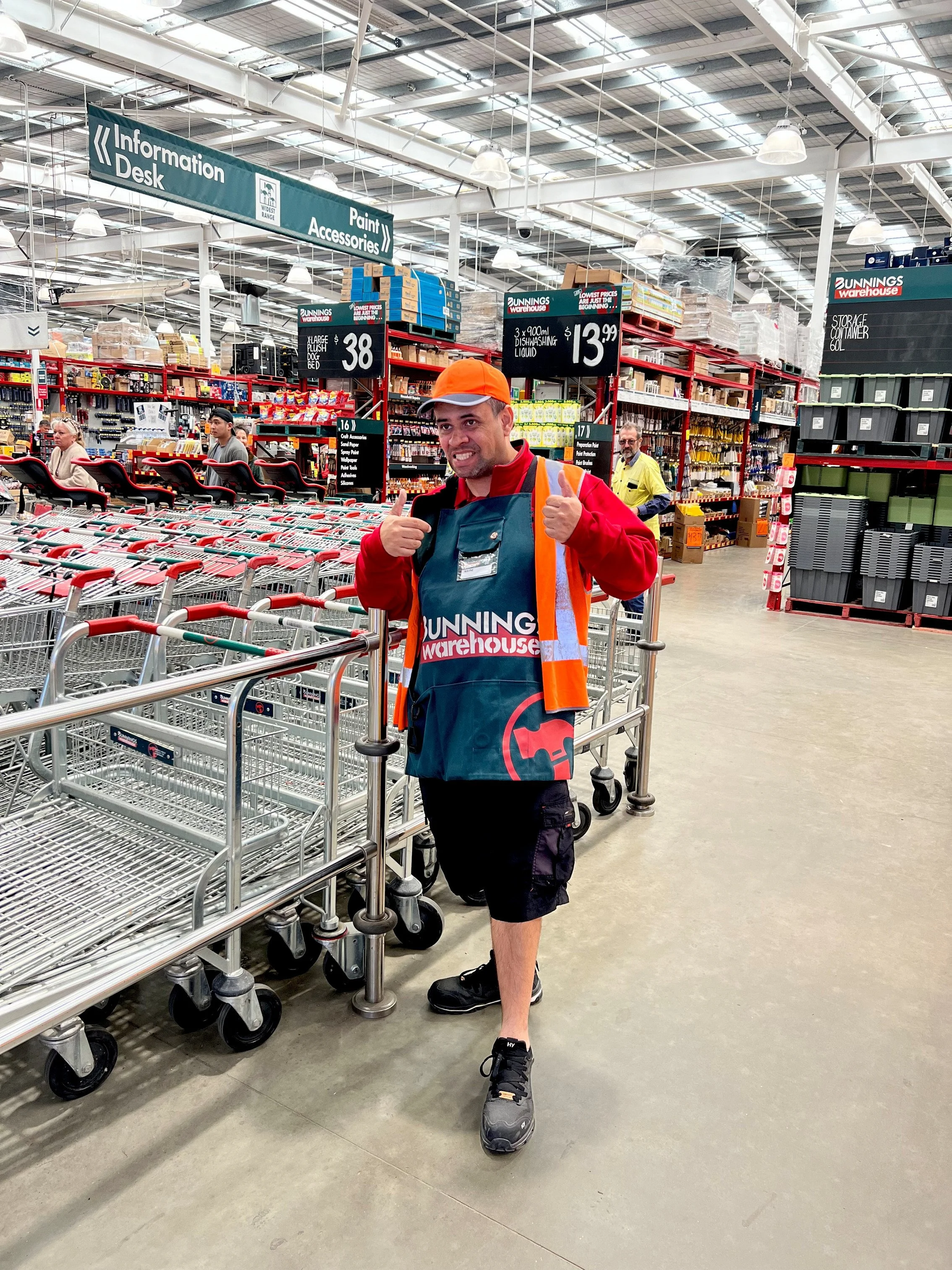 A man wearing a Bunnings Warehouse apron and orange safety vest giving a thumbs up inside a large hardware store.