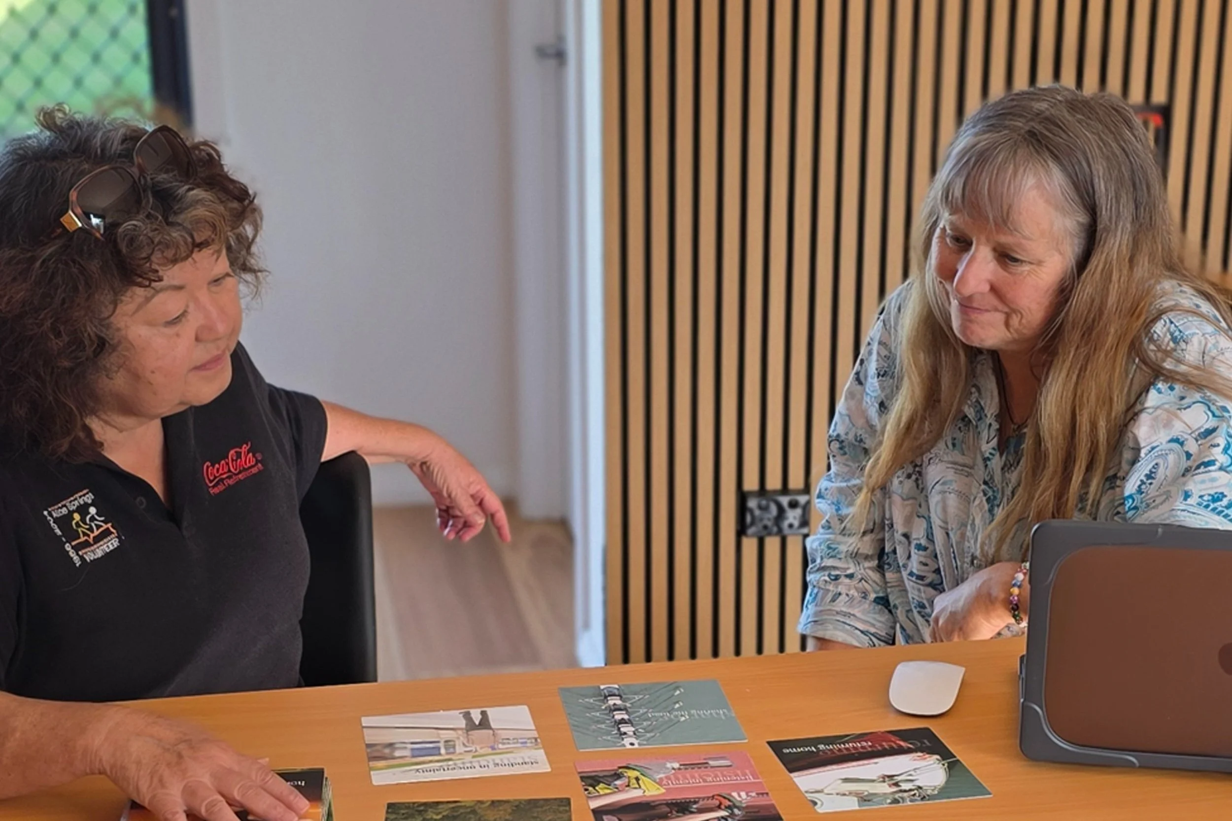 Two women sitting at a table with brochures and a laptop, engaged in conversation about NDIS services, in a room with wooden panel walls.