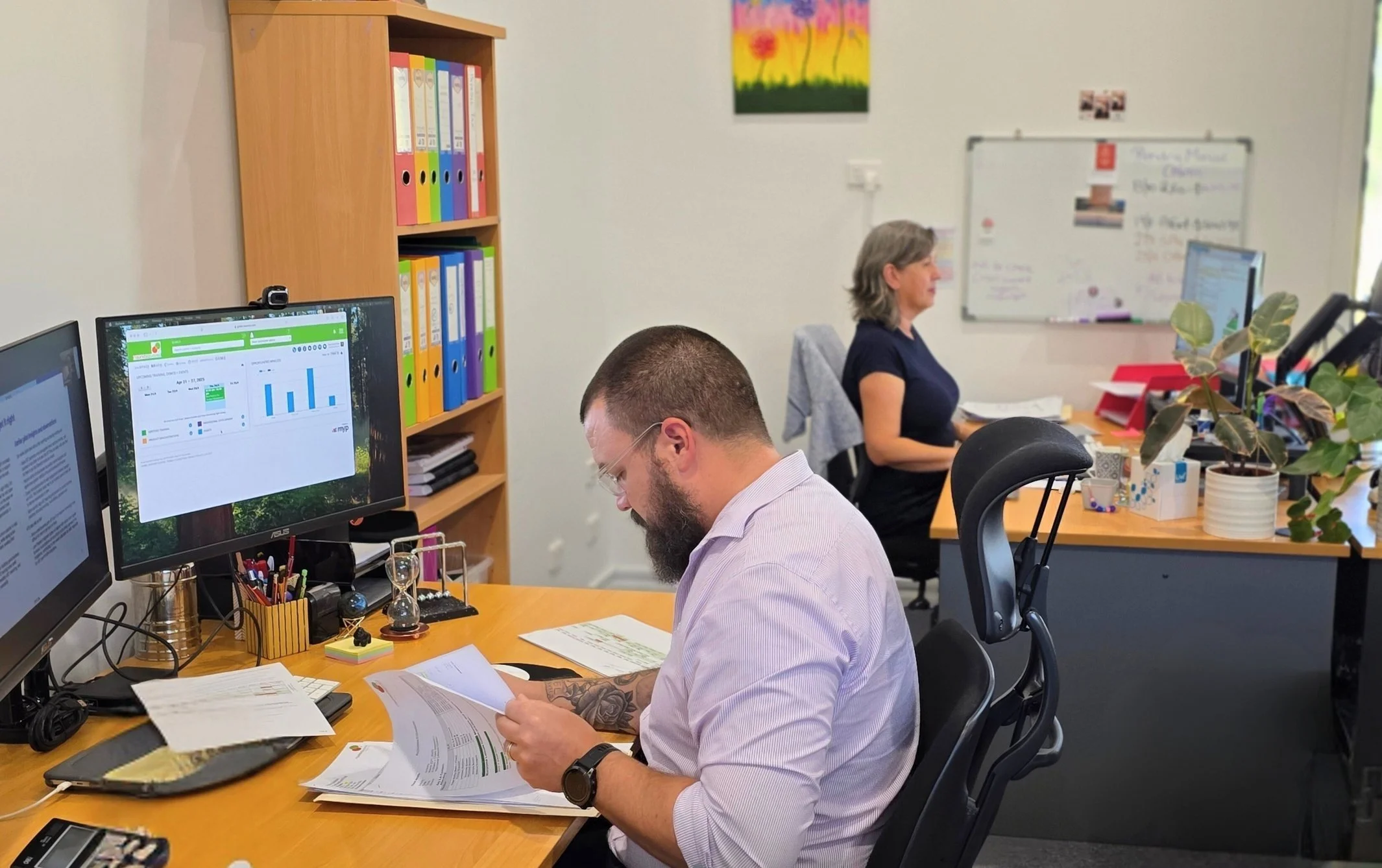 An office scene with two people working at desks. The man in the foreground has a beard, glasses, and tattoos, and is reading documents. The woman in the background has gray hair and is using a computer. There are multiple monitors, a whiteboard, bookshelf with colorful binders, and plants on the desks.