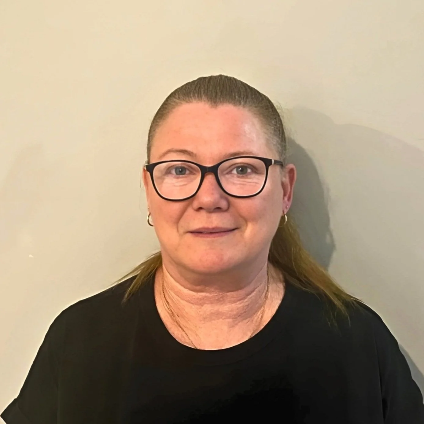 Image of Frances Minotte, Director wearing glasses, earrings, and long hair in a ponytail, wearing a black shirt, standing against a plain light-coloured wall.