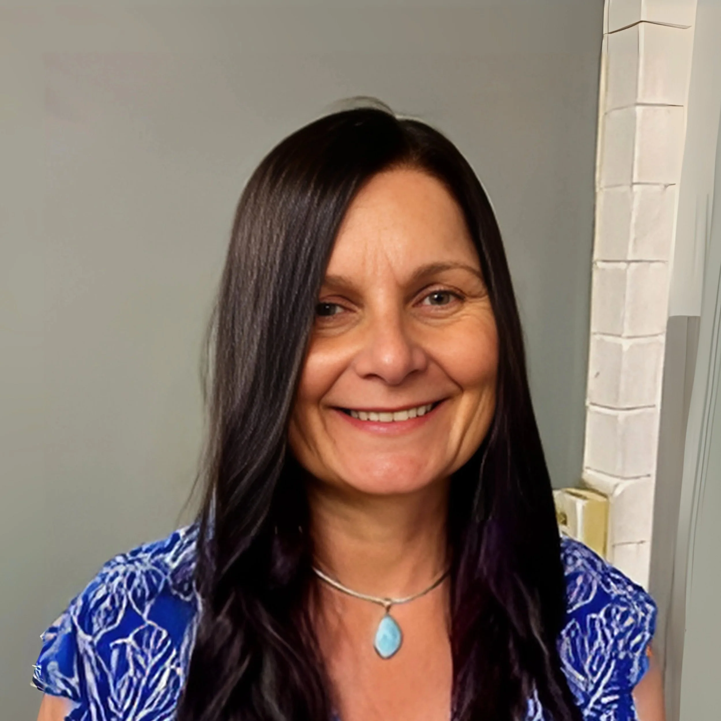 Image of Jodie Hoger, Director smiling at camera with long dark hair, wearing a blue patterned top and a silver necklace with a blue pendant.