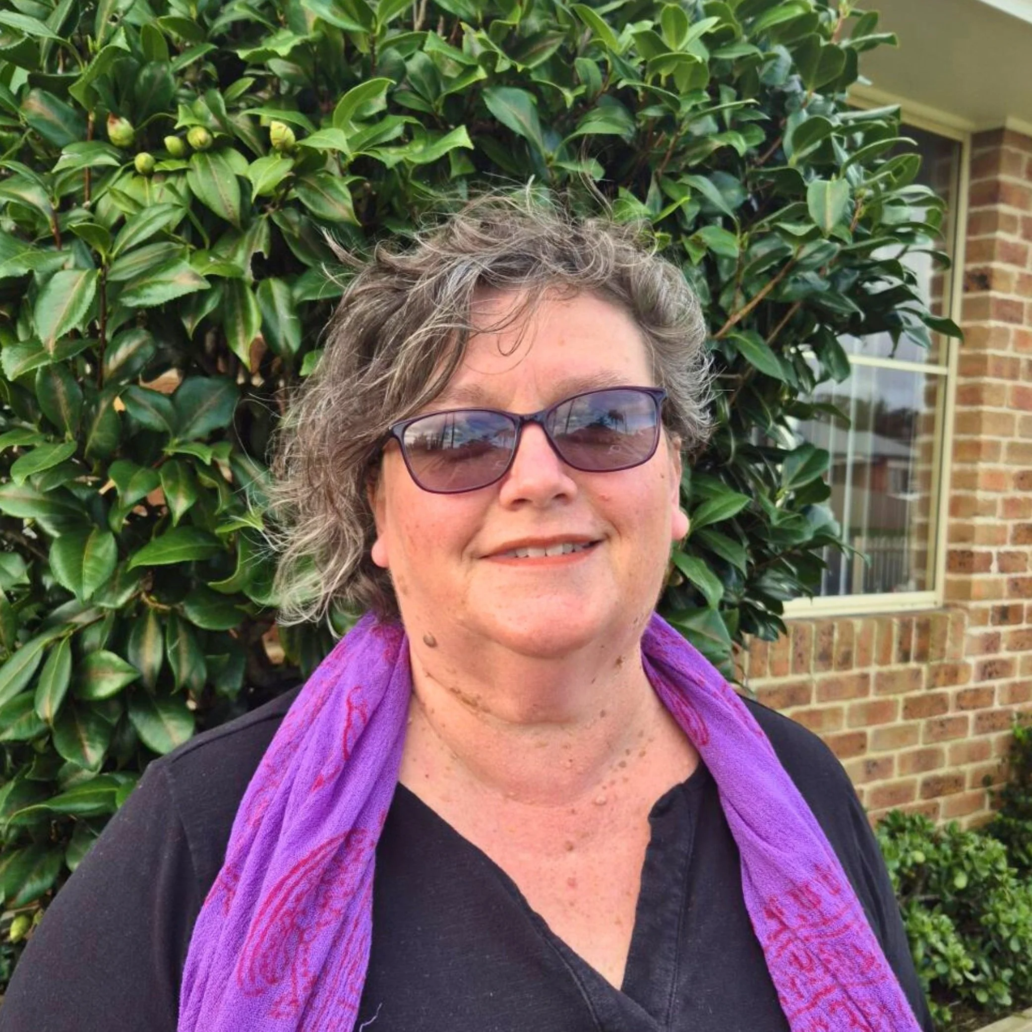 Image of Suzanne Kerslake, Director with curly short hair, wearing sunglasses, a purple scarf, and a black top, standing in front of a green bush and a brick house with windows.