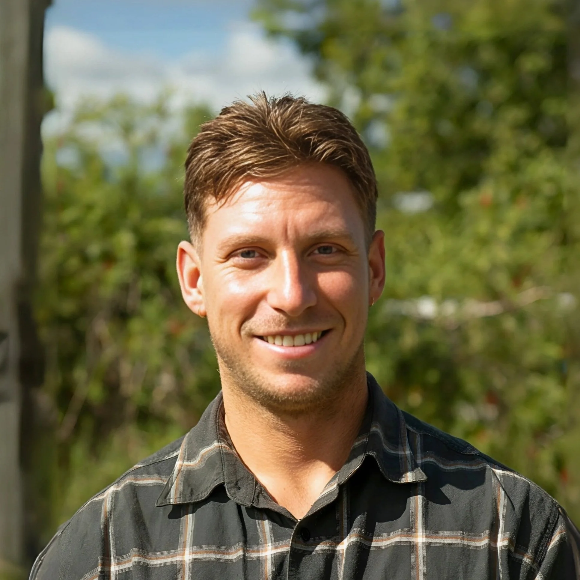 Image of Luke, Support Coordinator with short brown hair, wearing a plaid button up shirt, standing outdoors, smiling 