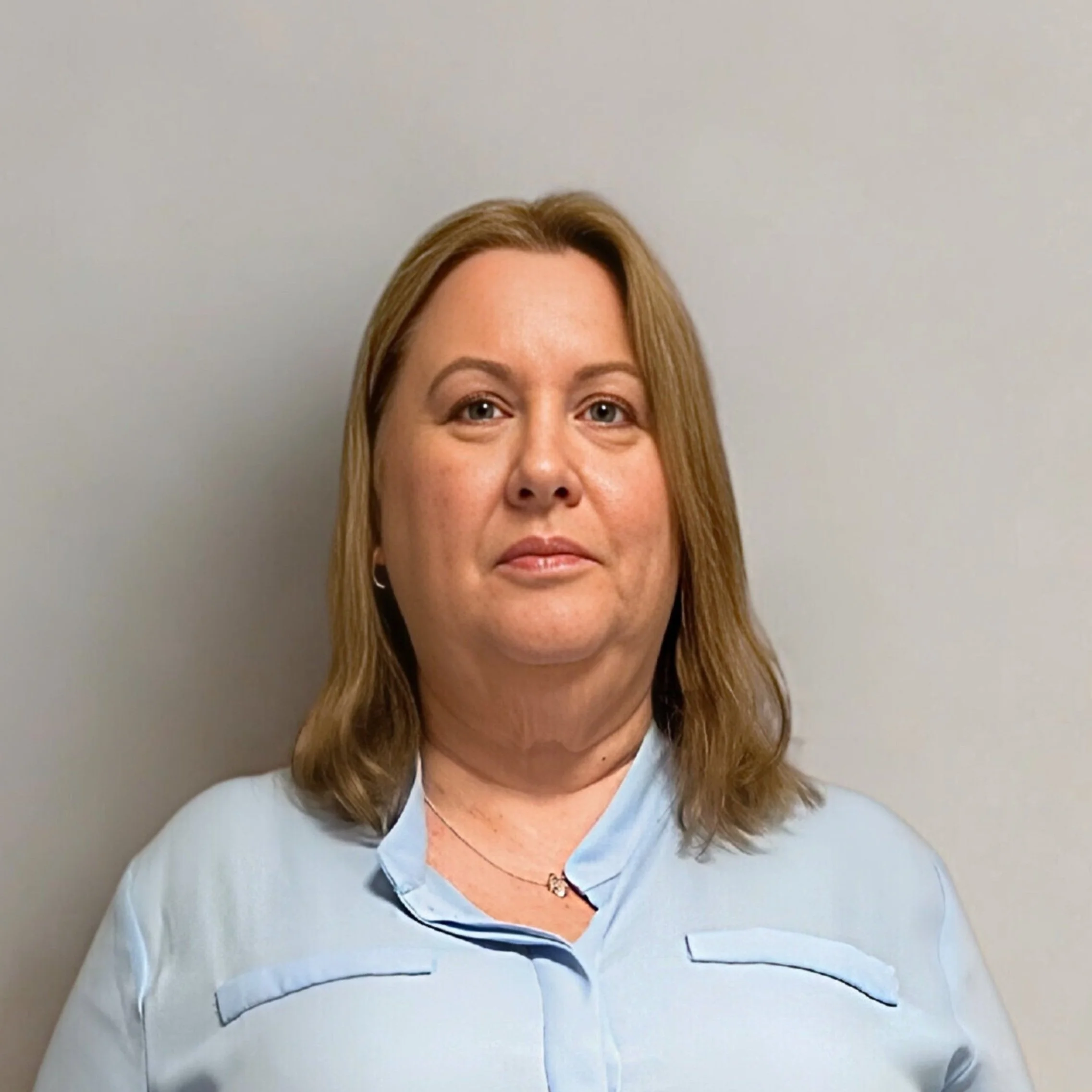 Image of Evelyn, Support Coordinator with shoulder-length light brown hair and fair skin, wearing a light blue collared shirt, standing against a plain off-white wall.