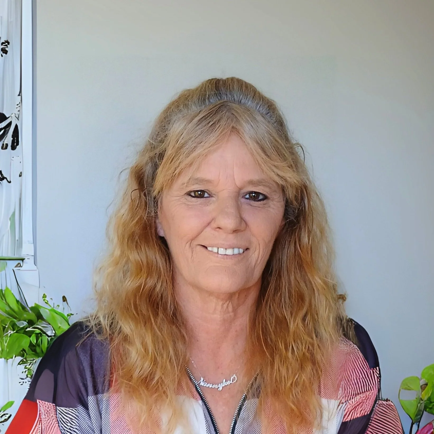 Image of Kelly, Support Coordinator, a woman with long, wavy blonde hair smiling in front of a plain background with green plants on the side.