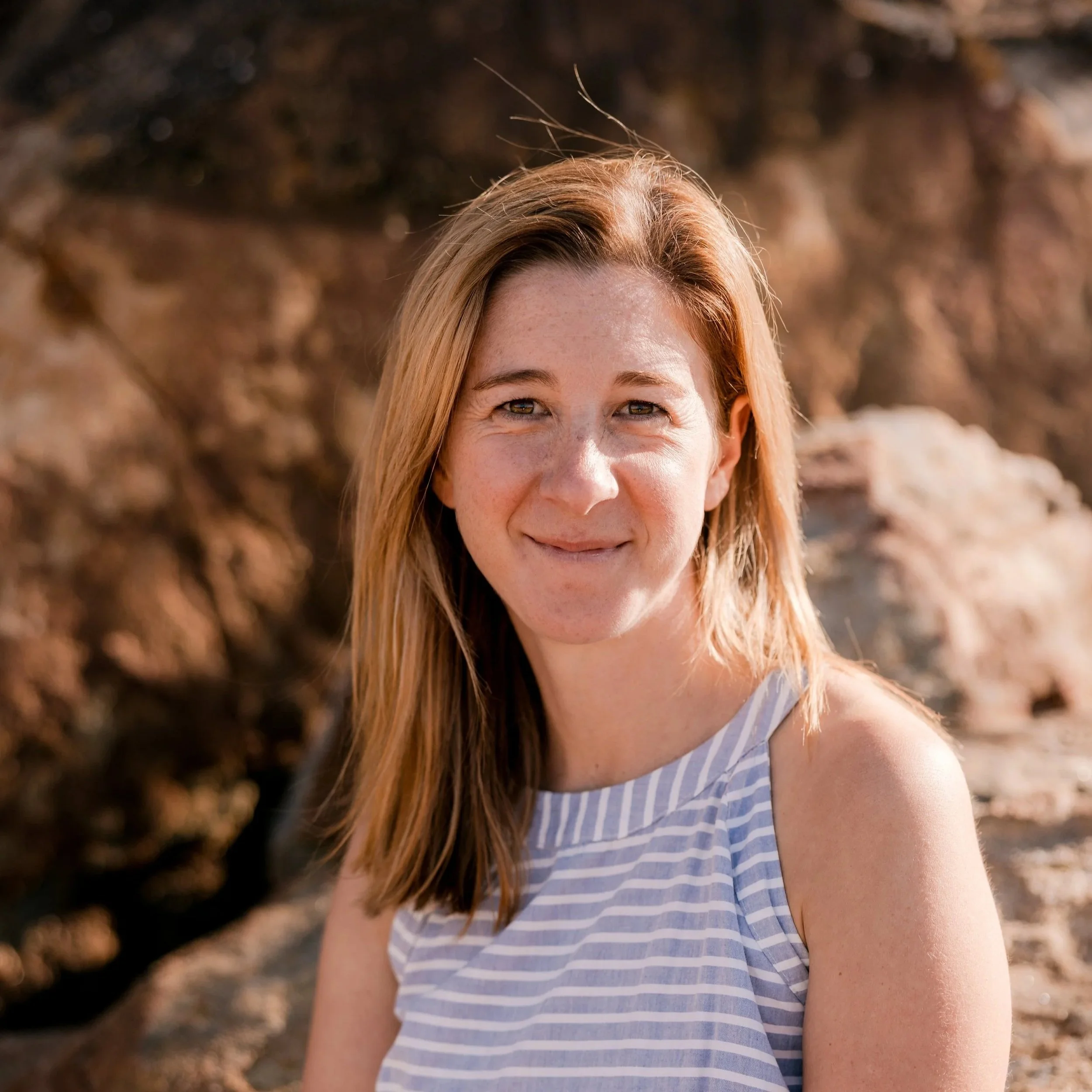 Image of Elise, Support Coordinator with shoulder-length curly blonde hair, wearing earrings and a black top, stands outdoors near a body of water with trees in the background, under a blue sky.