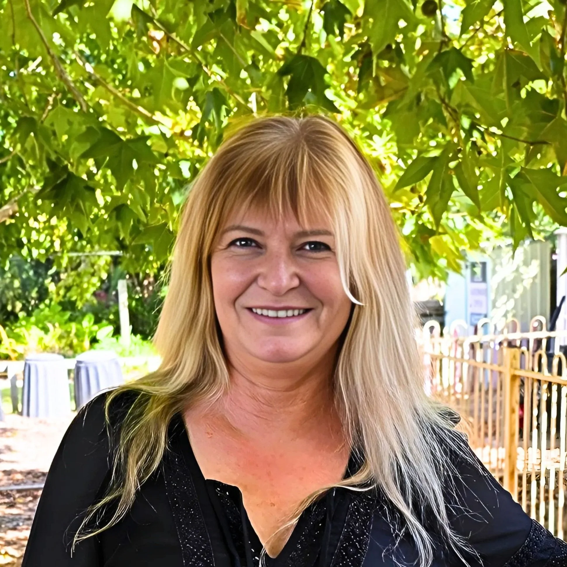 Image of Angela, Support Coordinator with long blonde hair smiling outdoors with green leaves and trees in the background.