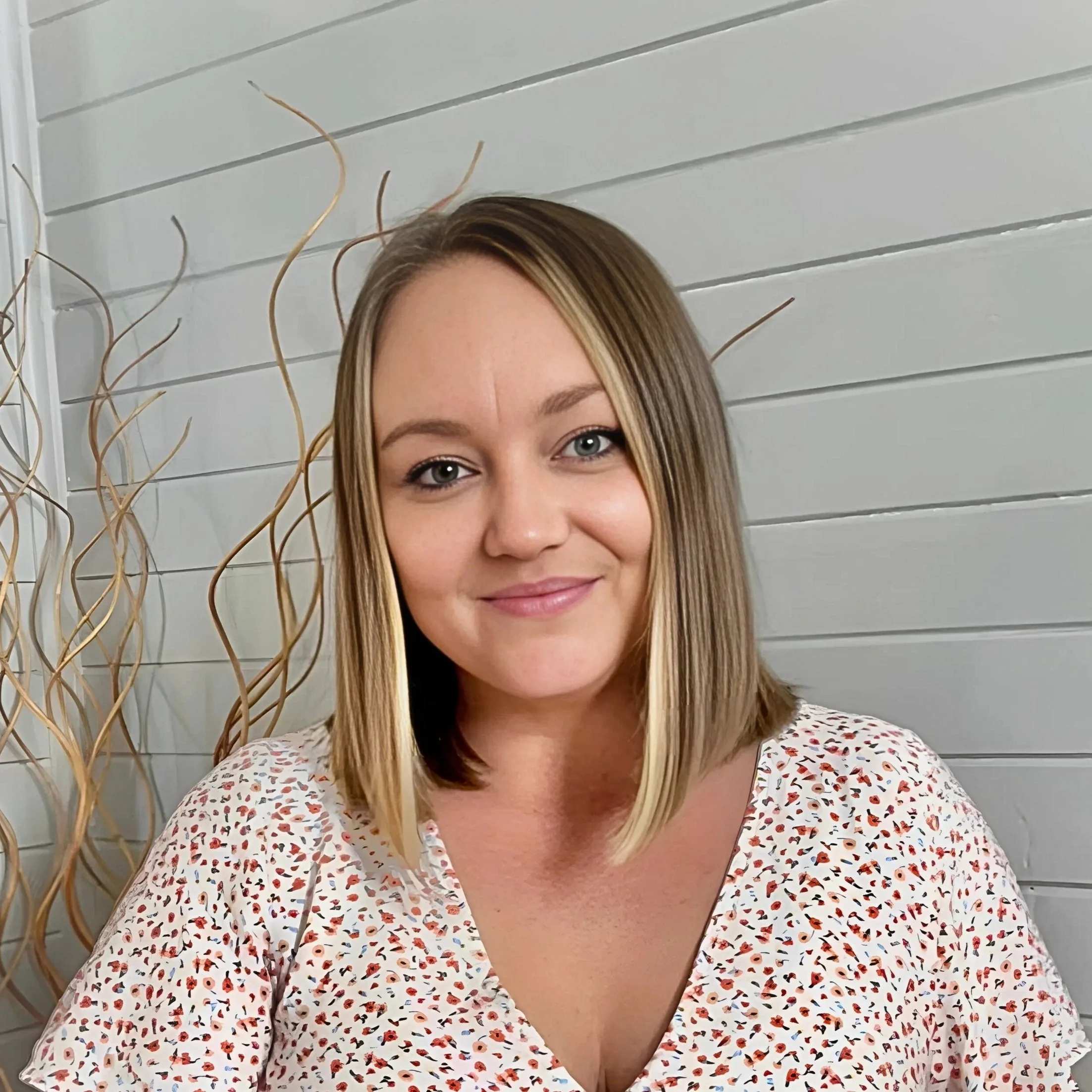 Image of Alana, Support Coordinator with shoulder-length blonde hair, blue eyes, and a subtle smile, wearing a white blouse with a red and black floral pattern, standing against a white paneled wall with decorative wooden branches.