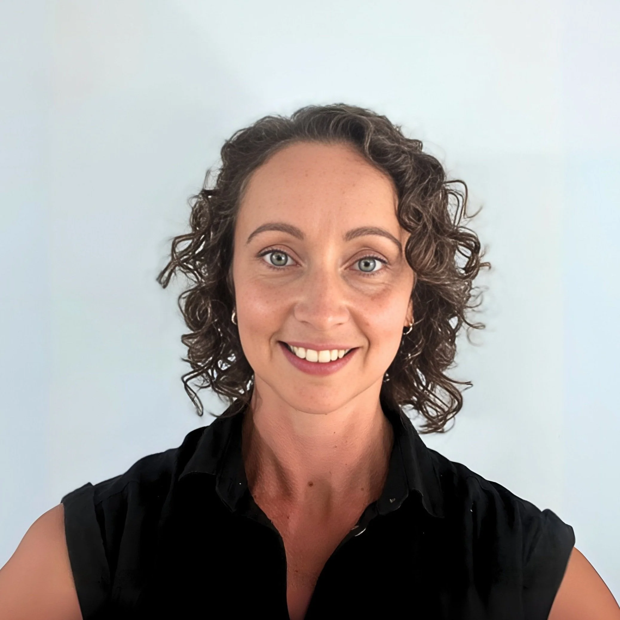 Image of Belinda, Support Coordinator with curly brown hair, light blue eyes, wearing a black sleeveless shirt, smiling against a plain white background.