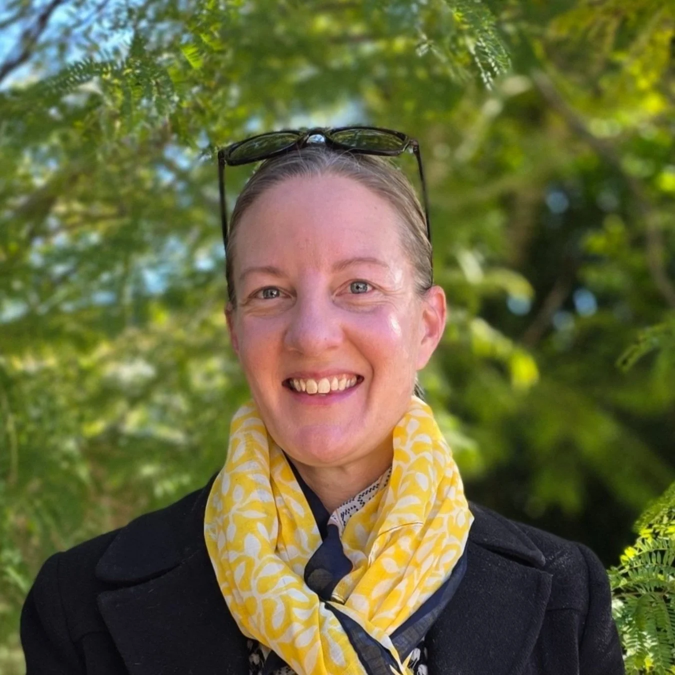 Image of Claire, Support Coordinator with blonde hair pulled back, wearing sunglasses on her head, a black jacket, and a yellow patterned scarf, standing outdoors with green foliage in the background.