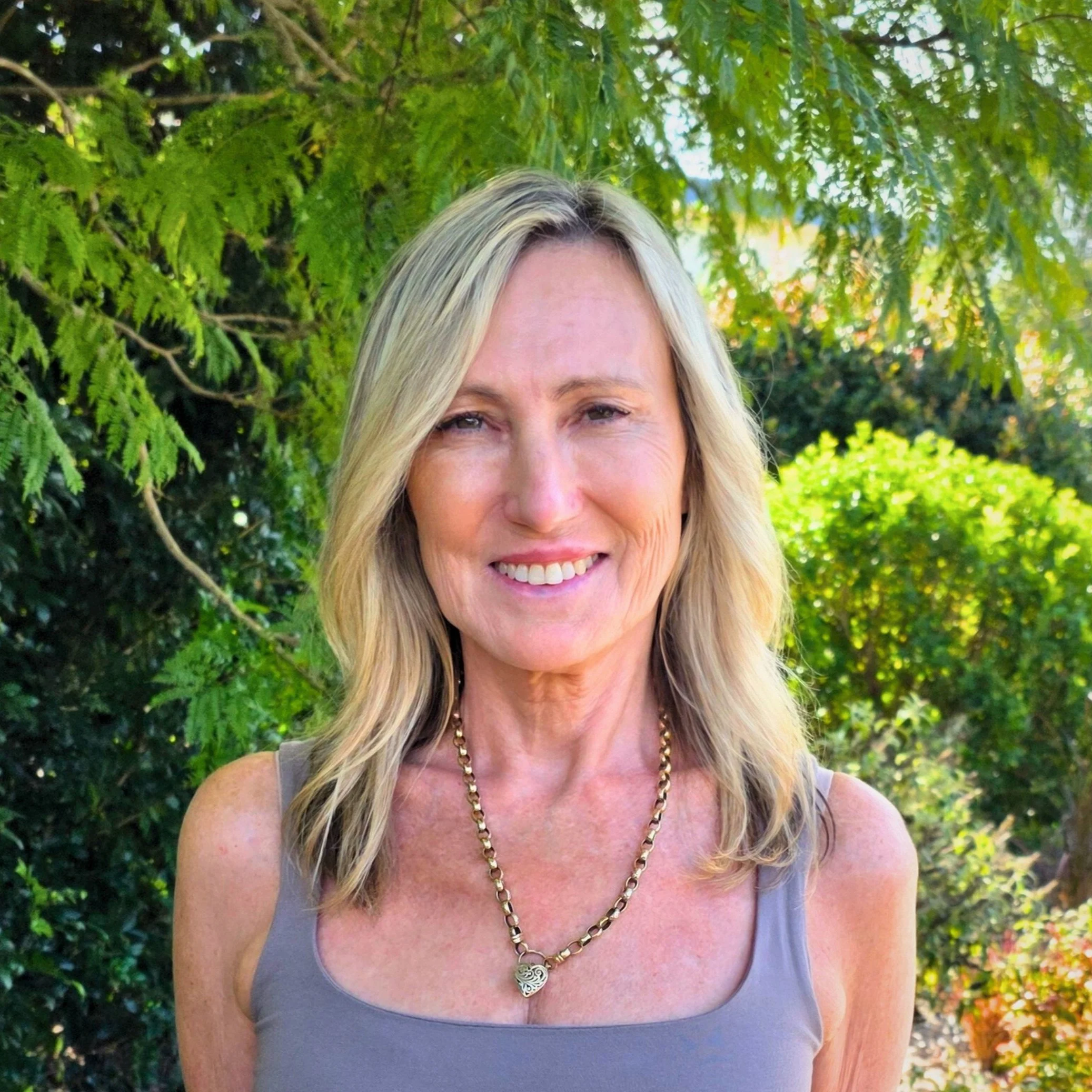 Image of Karen, Support Coordinator with blonde hair wearing a gray sleeveless top and a gold chain necklace standing outdoors in front of green trees and bushes.