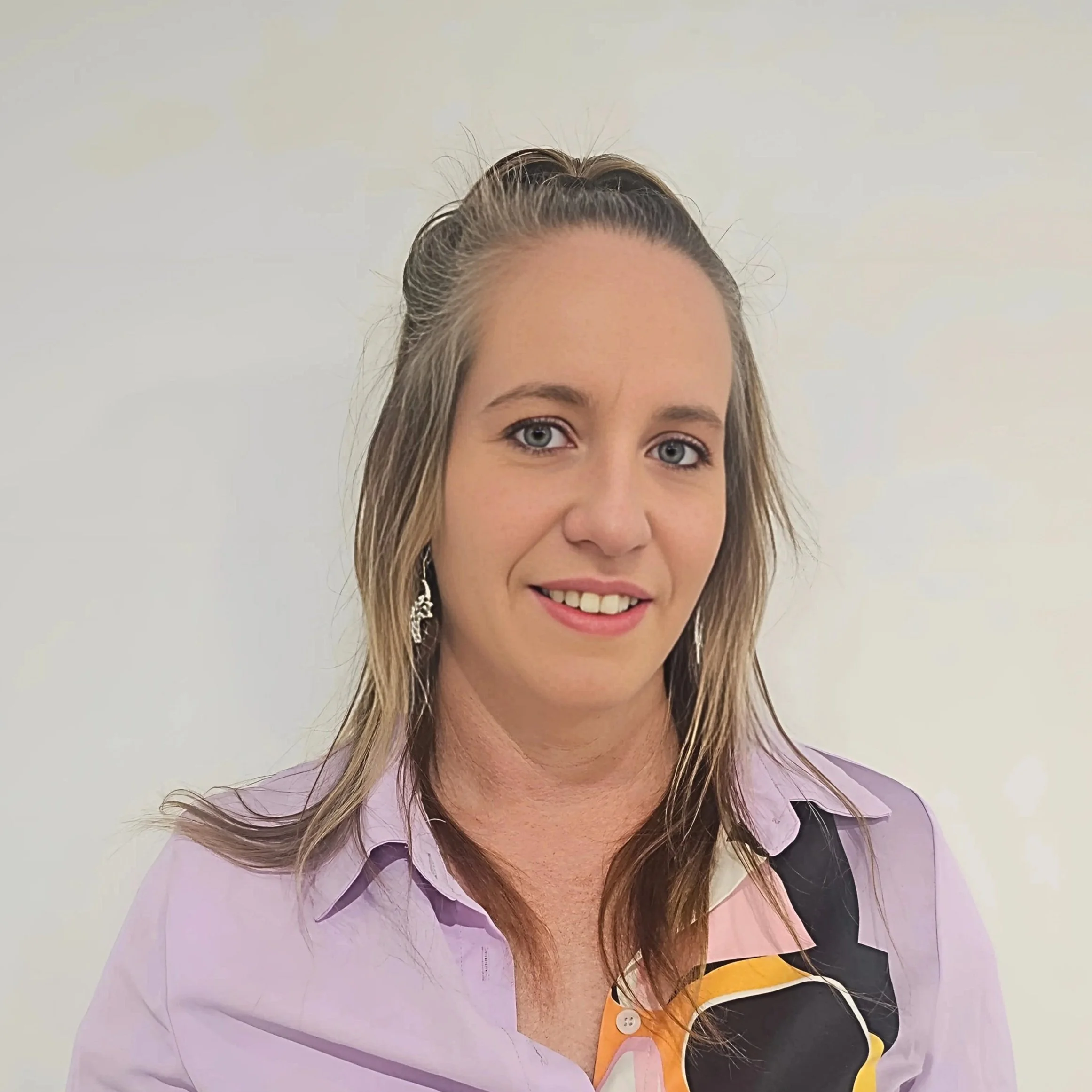 Image of Kristy M, Support Coordinator with long, light brown hair partially pulled back, wearing a light purple button-up shirt, earrings, and smiling against a plain white background.