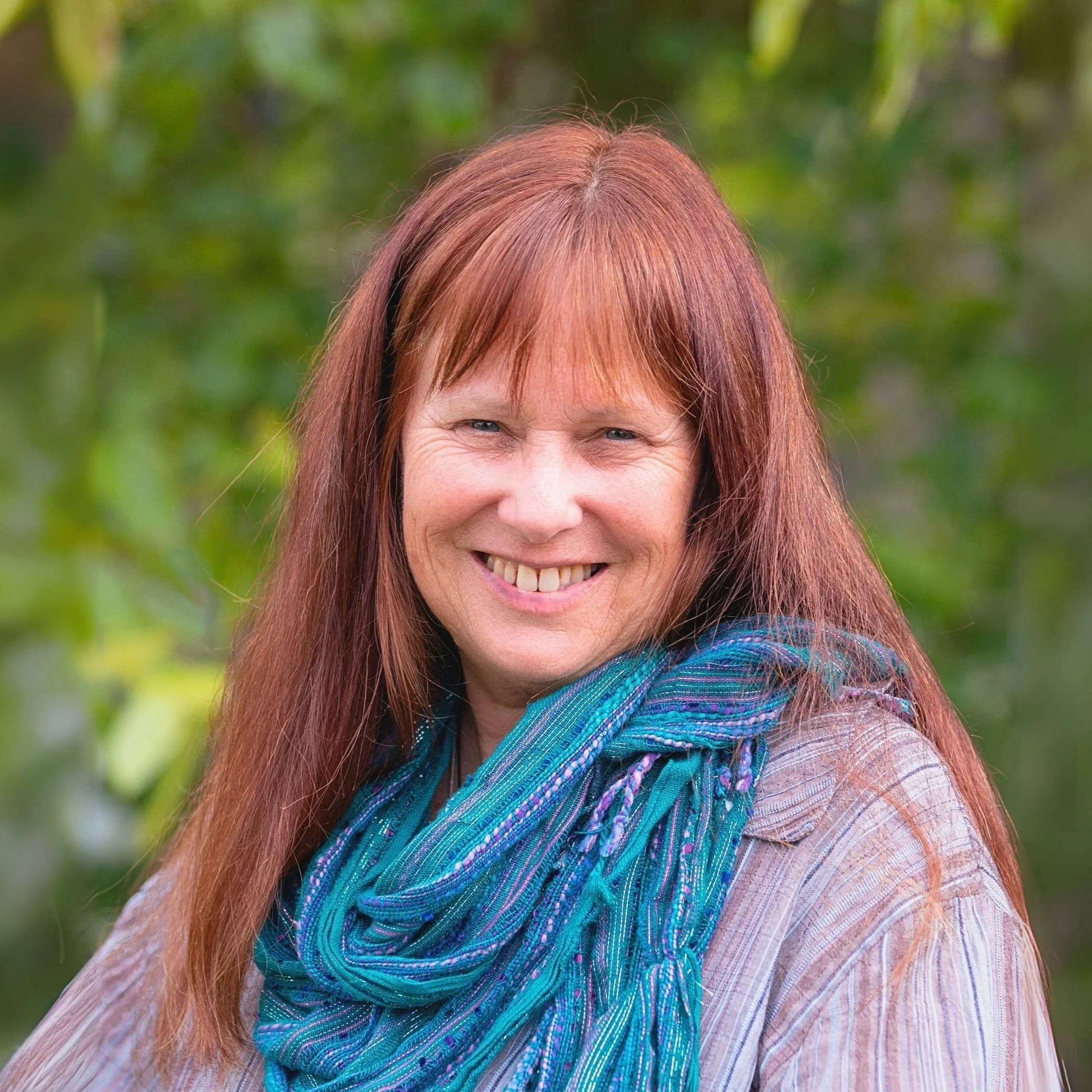 Image of Sam, Support Coordinator A woman with long red hair, blue eyes, and a bright smile, wearing a blue scarf and a light-colored top, outdoors against a green, blurred foliage background.